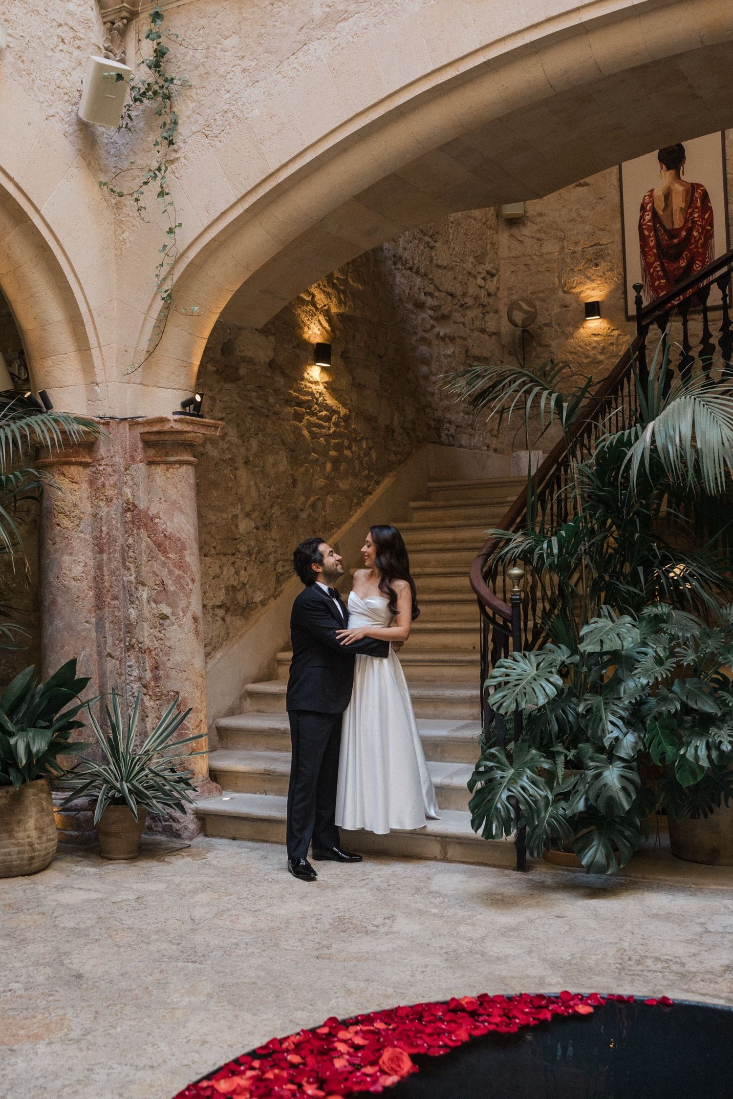 Couple shares intimate moment in historic stone courtyard with curved staircase and tropical plants near red rose petals.
