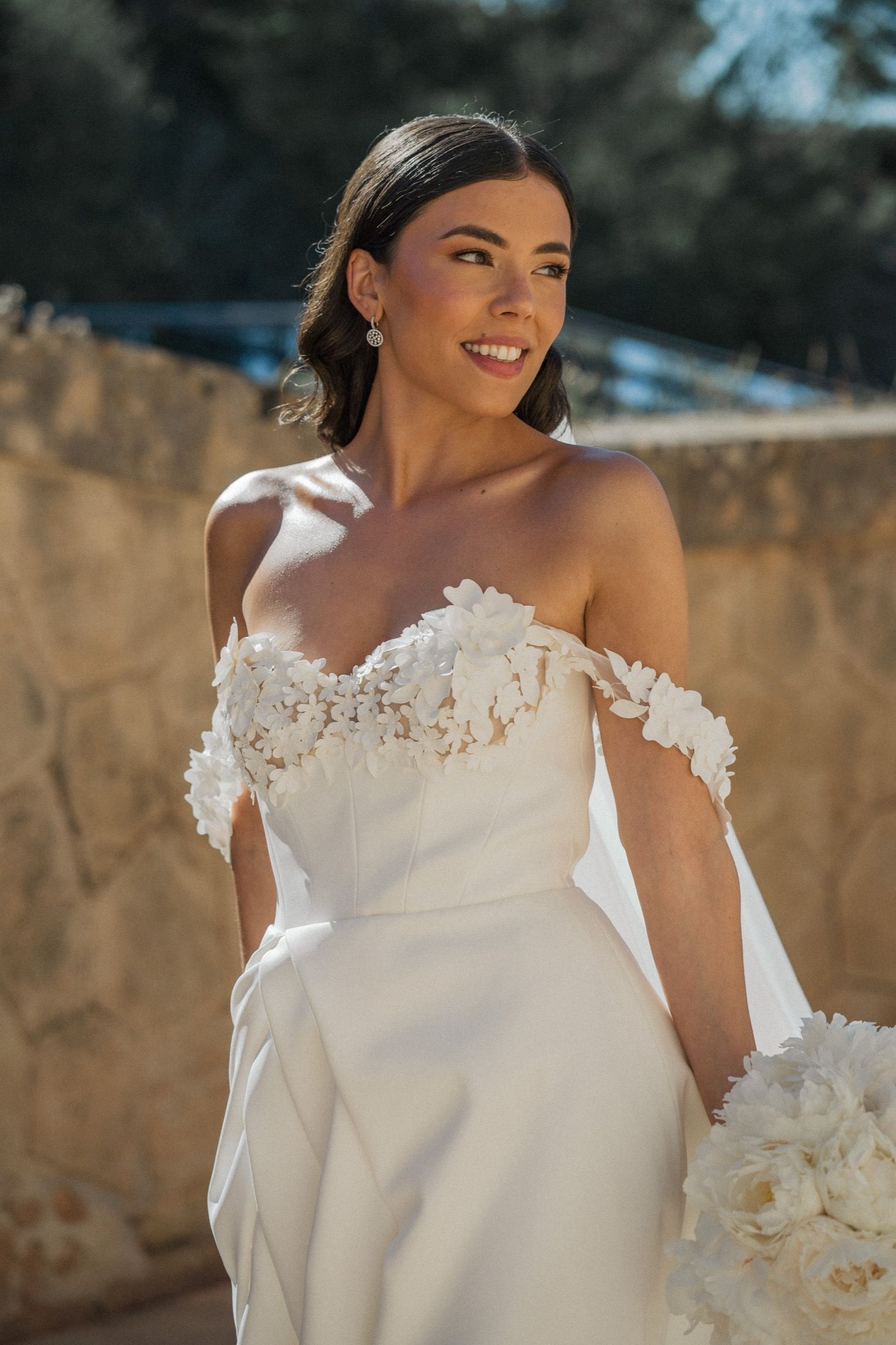 Smiling bride in off-shoulder white floral wedding gown holding bouquet outdoors in warm sunlight.
