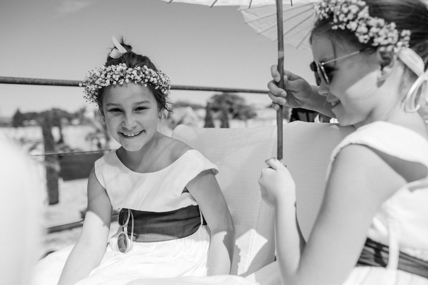 Two young flower girls in white dresses and floral crowns smile together at an outdoor wedding celebration.