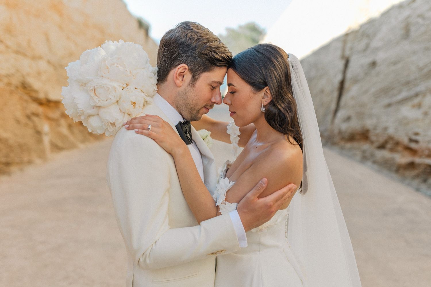 A bride and groom share an intimate moment outdoors, with the bride holding a white bouquet and wearing a veil.
