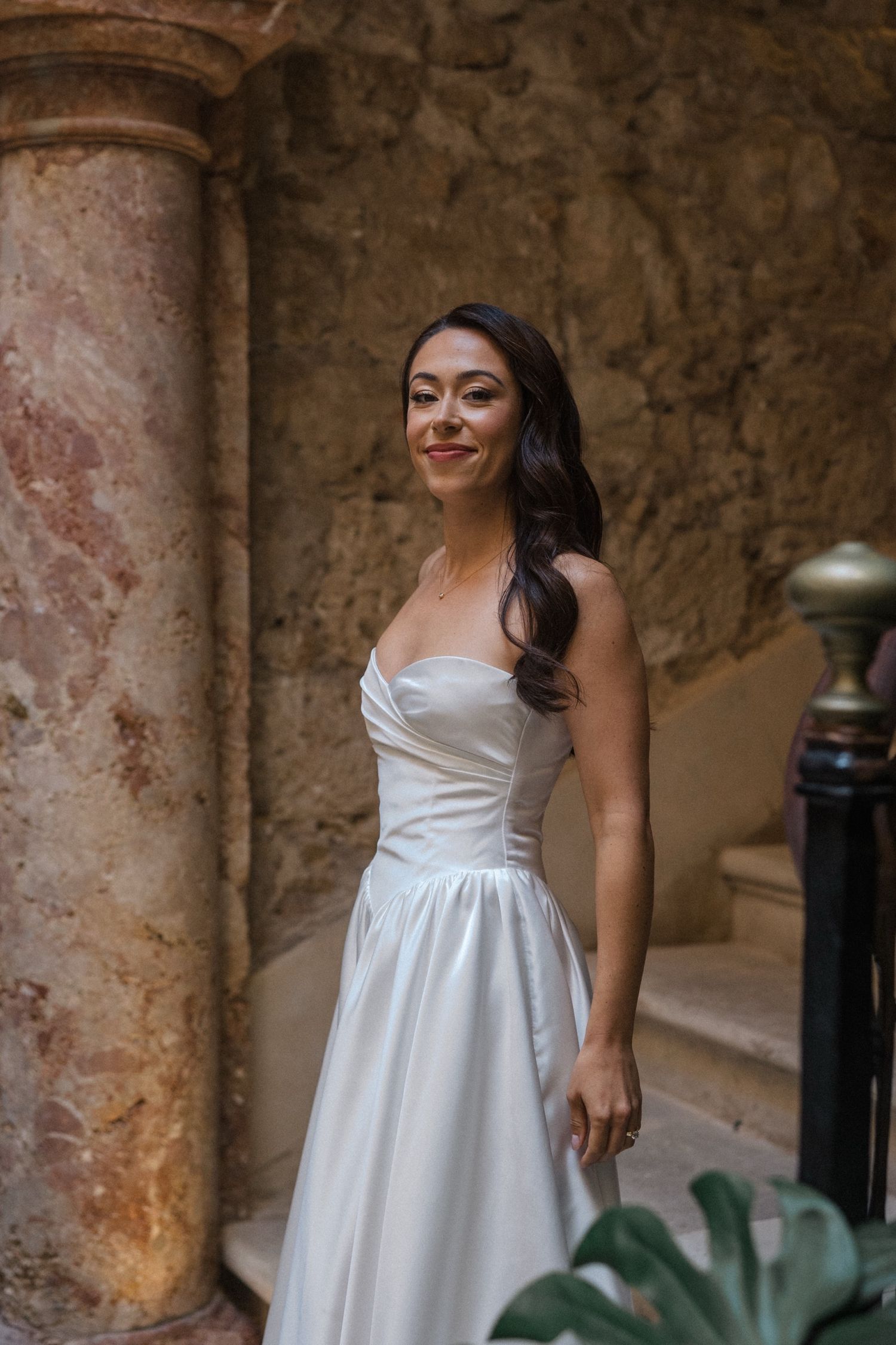 A bride in a white strapless wedding dress smiles while standing beside an ornate stone column in a rustic setting.