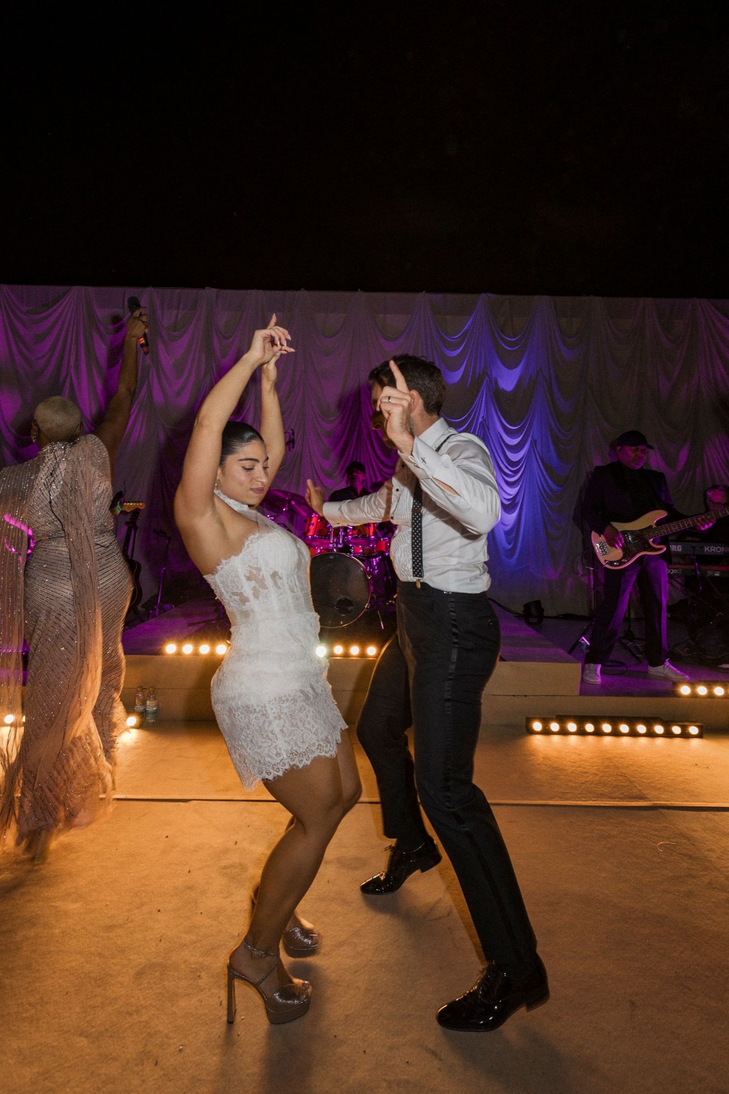 Dancers perform swing dance moves on a dimly lit stage with purple lighting and stage lights in the background.