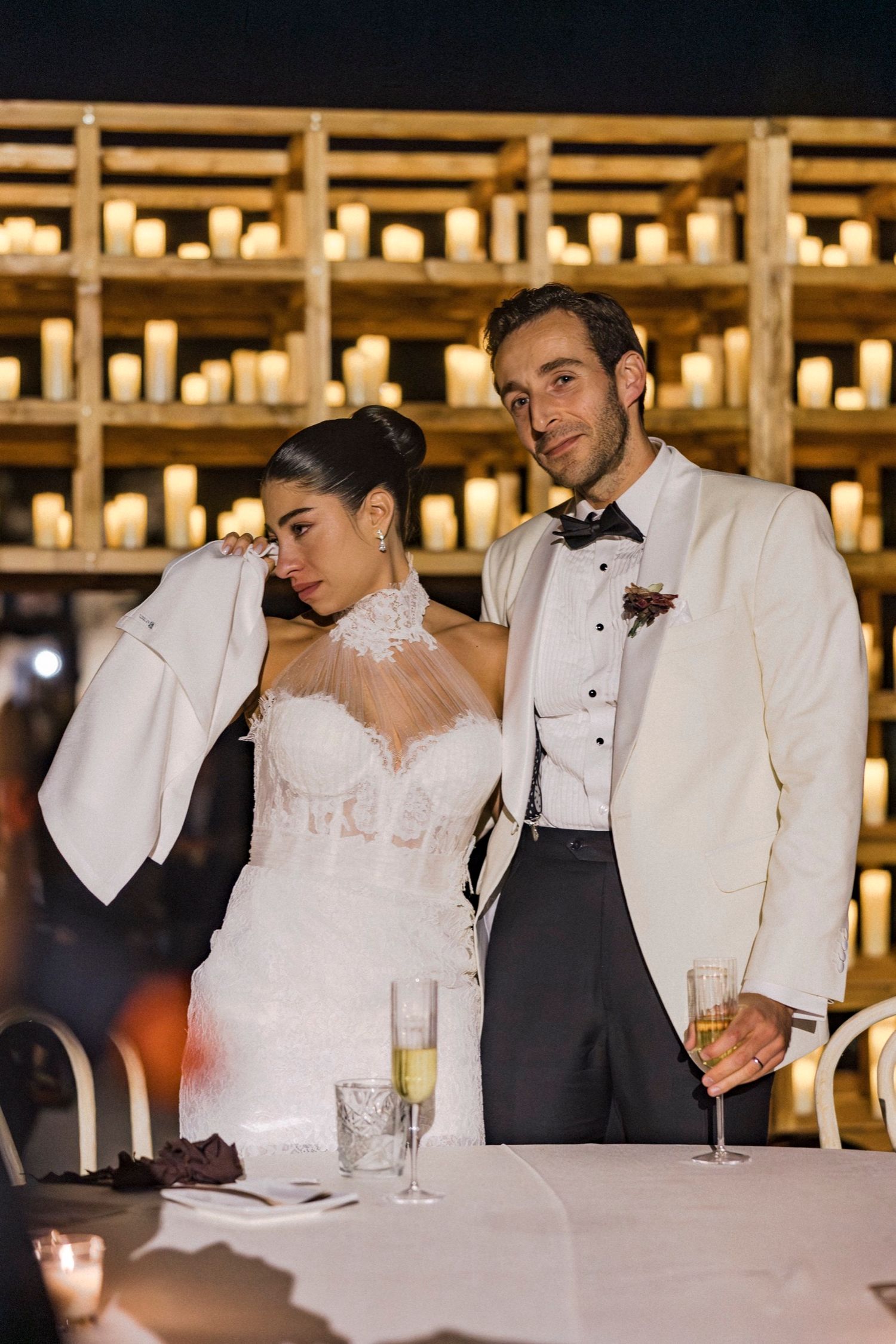A couple in elegant wedding attire share a moment at their reception with candlelit shelves in the background.