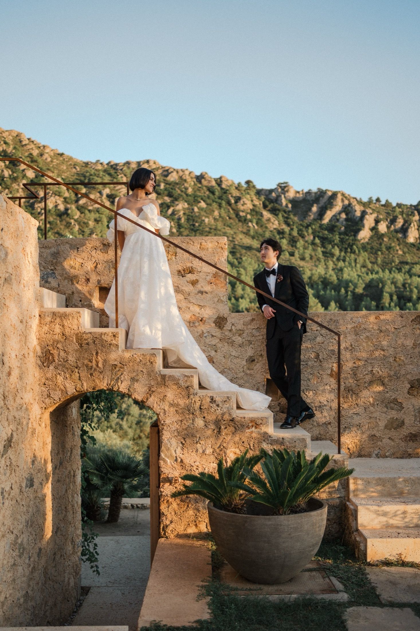 A bride in white dress and groom in tuxedo stand on stone steps with mountains in background.