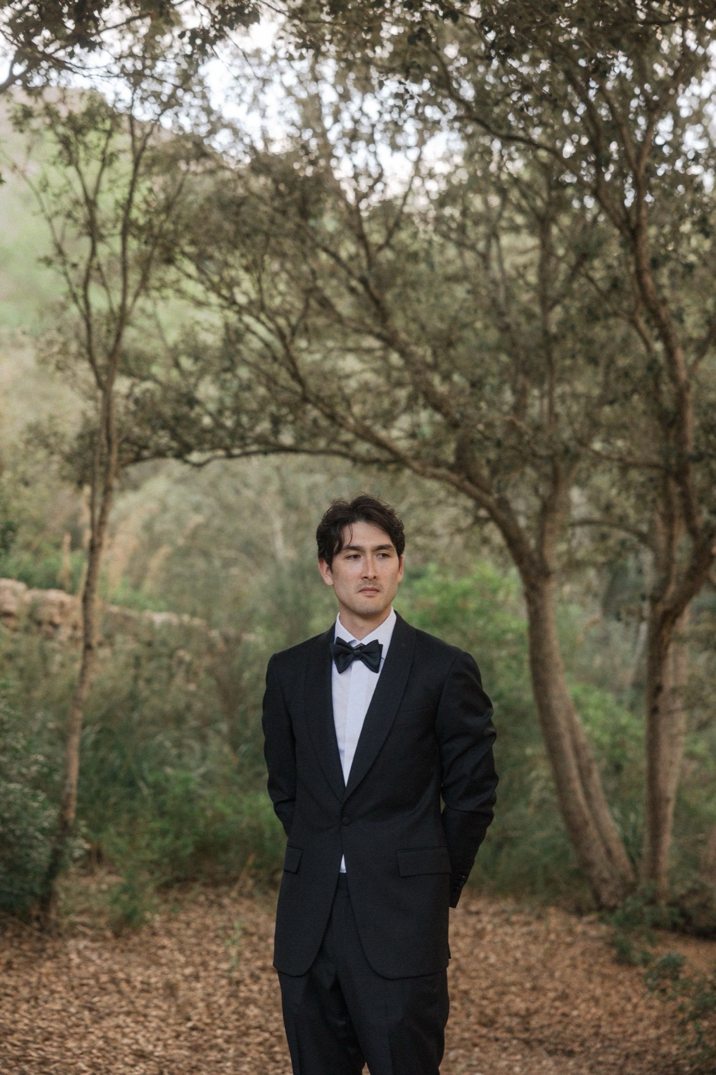 Person in formal black tuxedo standing in wooded area with fallen leaves on ground.