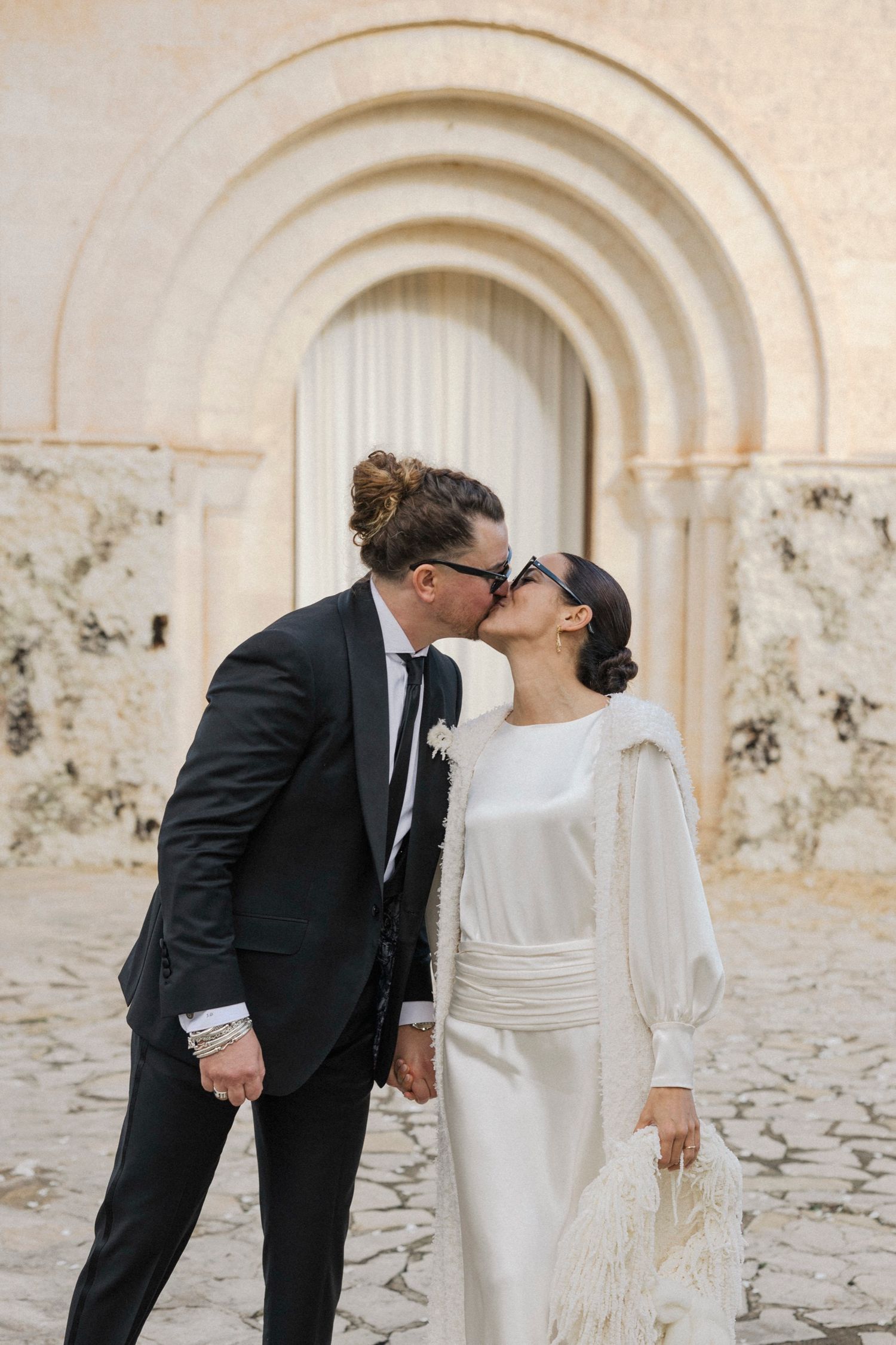 Romantic wedding kiss in front of an ornate arched doorway with stone walls.