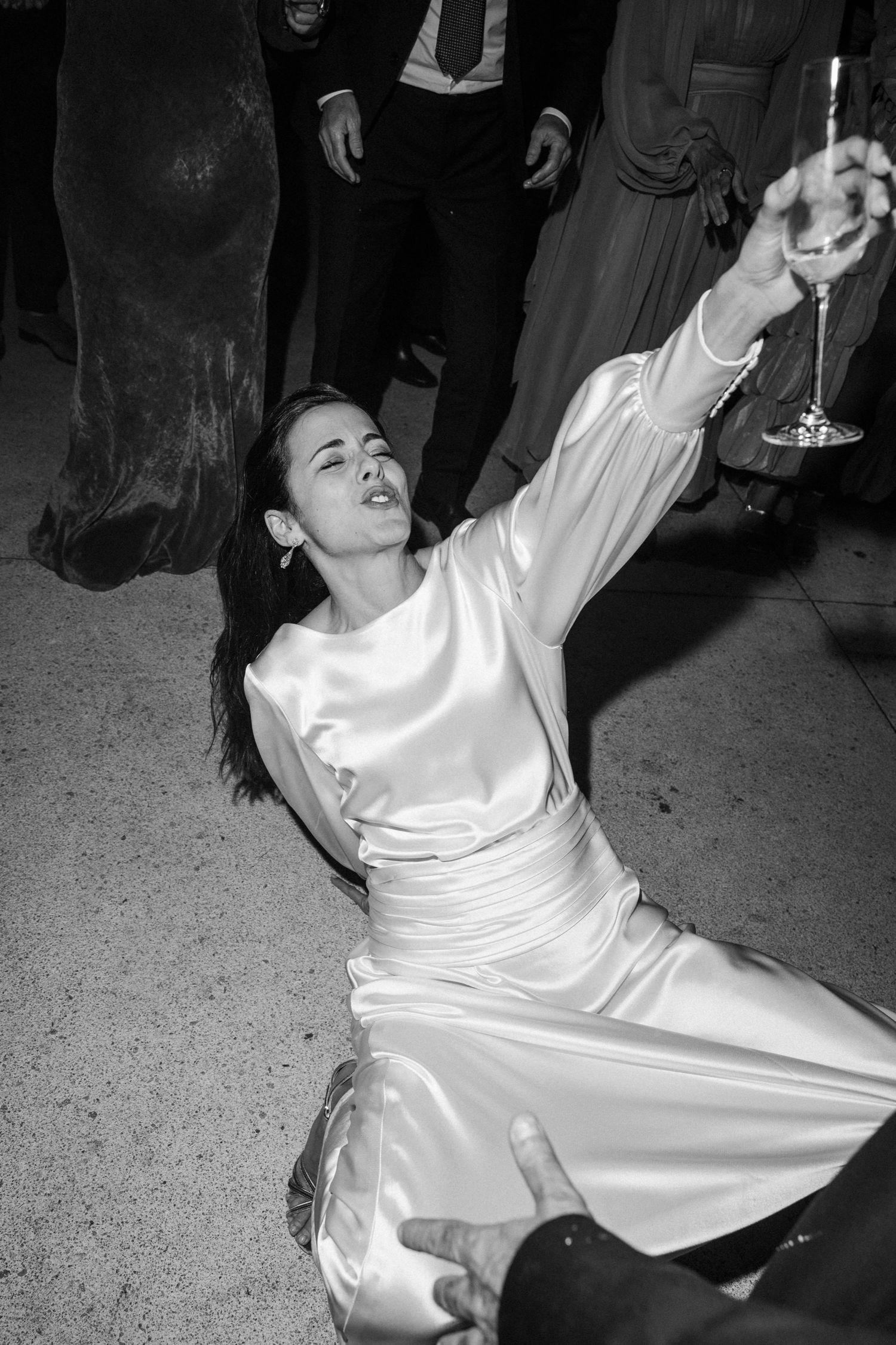 A person in a white satin dress sits on the floor while raising a champagne glass in celebration.