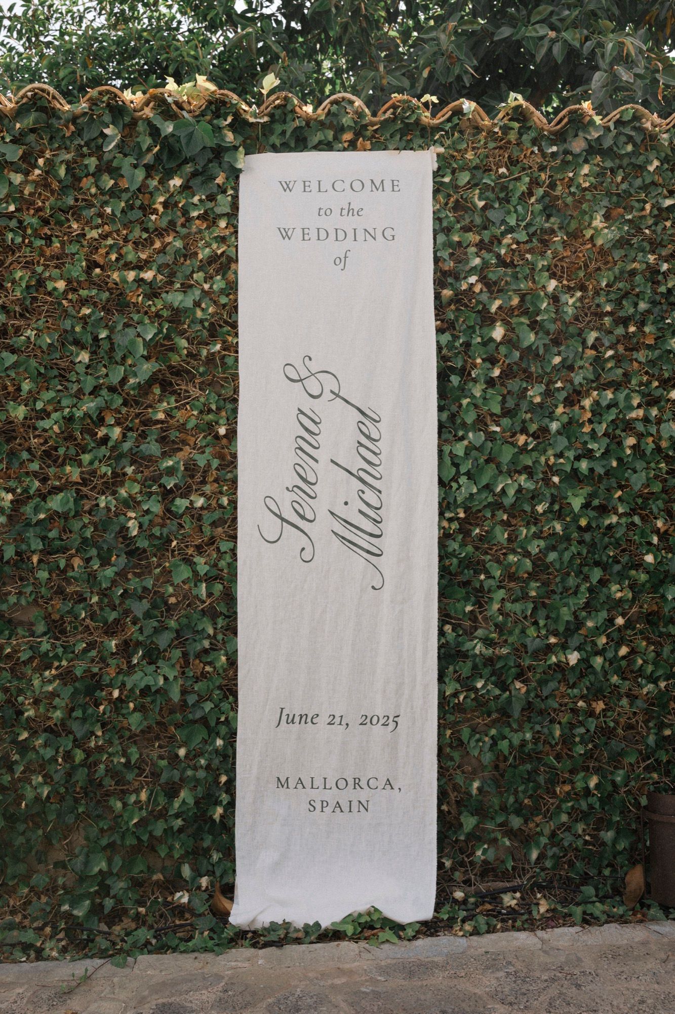 Elegant wedding welcome sign against green hedge wall displaying event details in Mallorca, Spain.