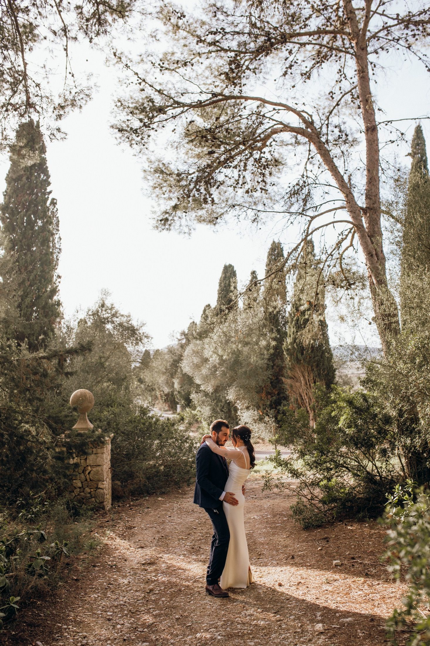 A romantic couple embraces on a sunlit path lined with tall cypress and pine trees in a Mediterranean setting.