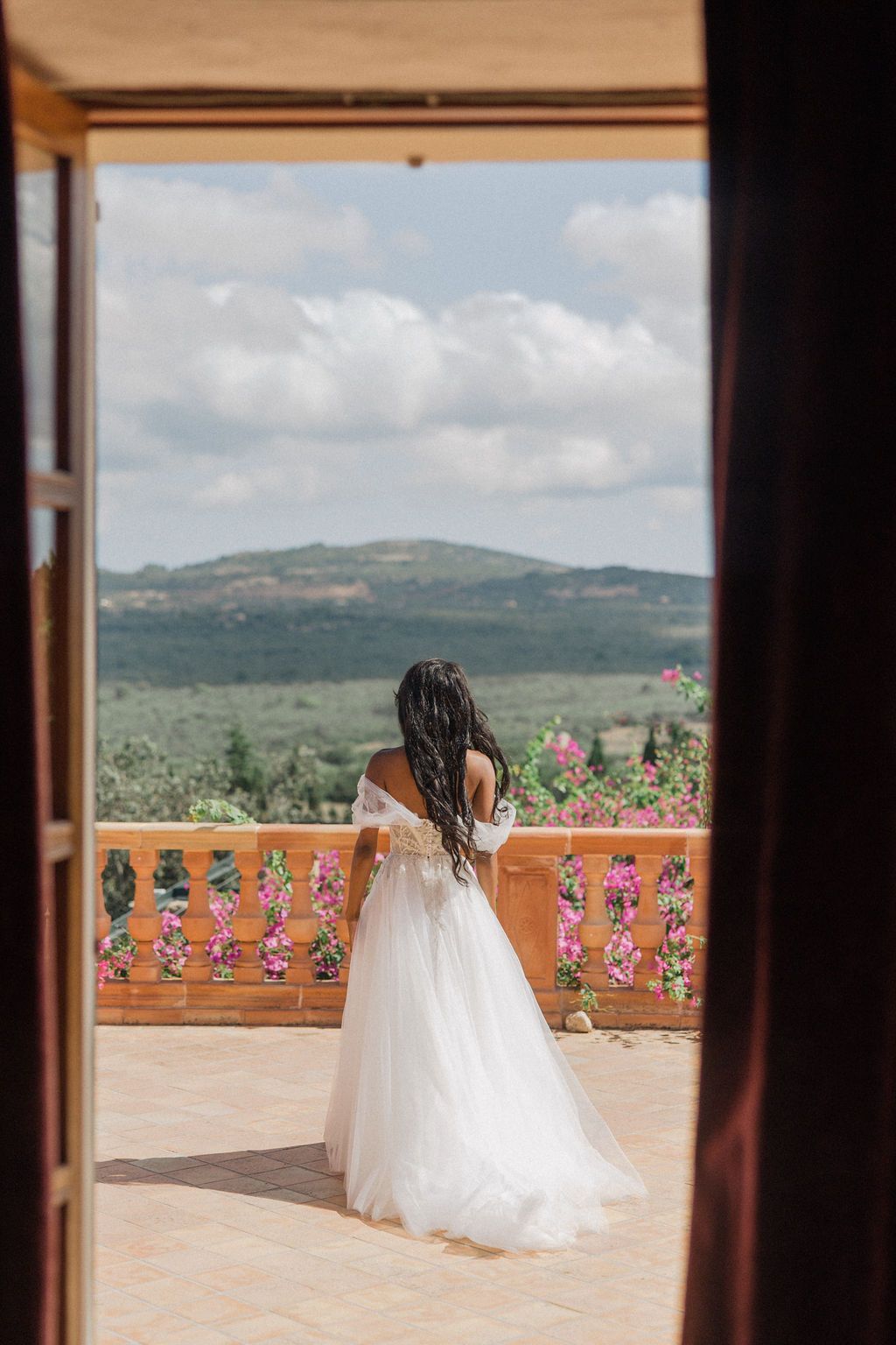 Wedding dress captured through doorway on terrace with mountain vista and pink flowers.