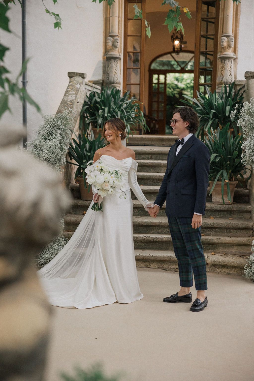 A couple stands on stone steps, the bride in a flowing white gown and groom in plaid pants.
