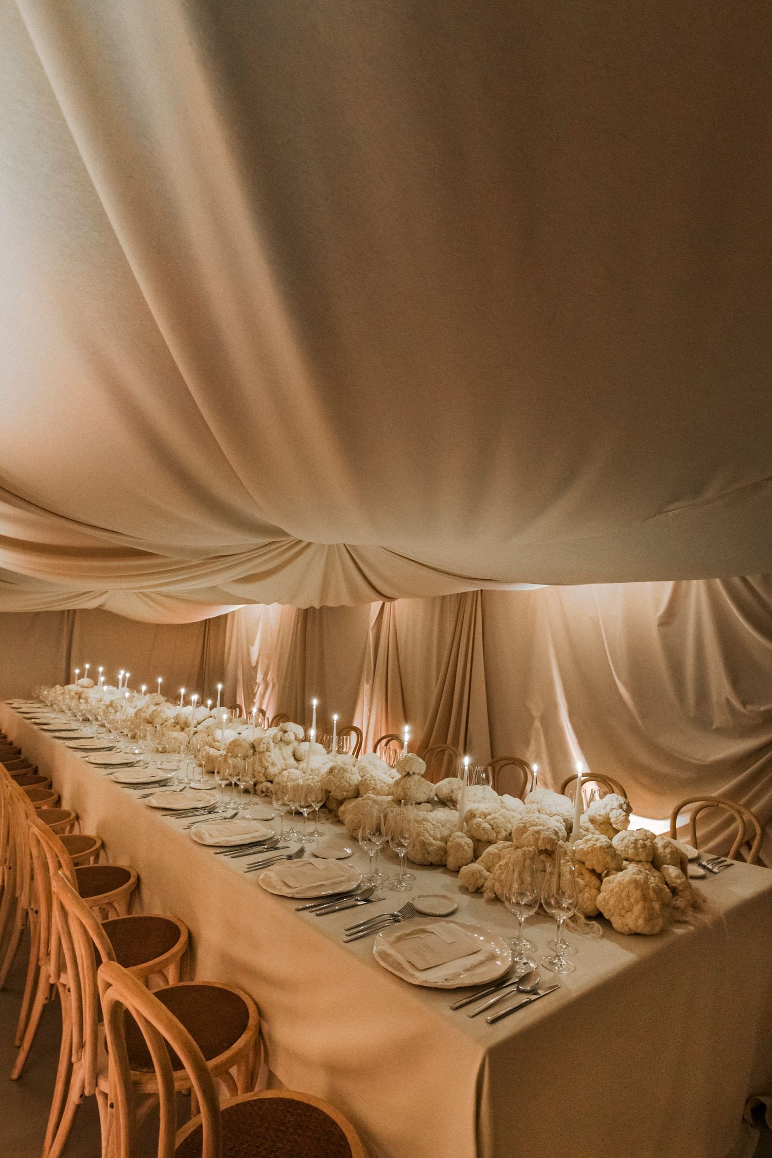Long formal dining table with white floral centerpieces and taper candles under draped ceiling.