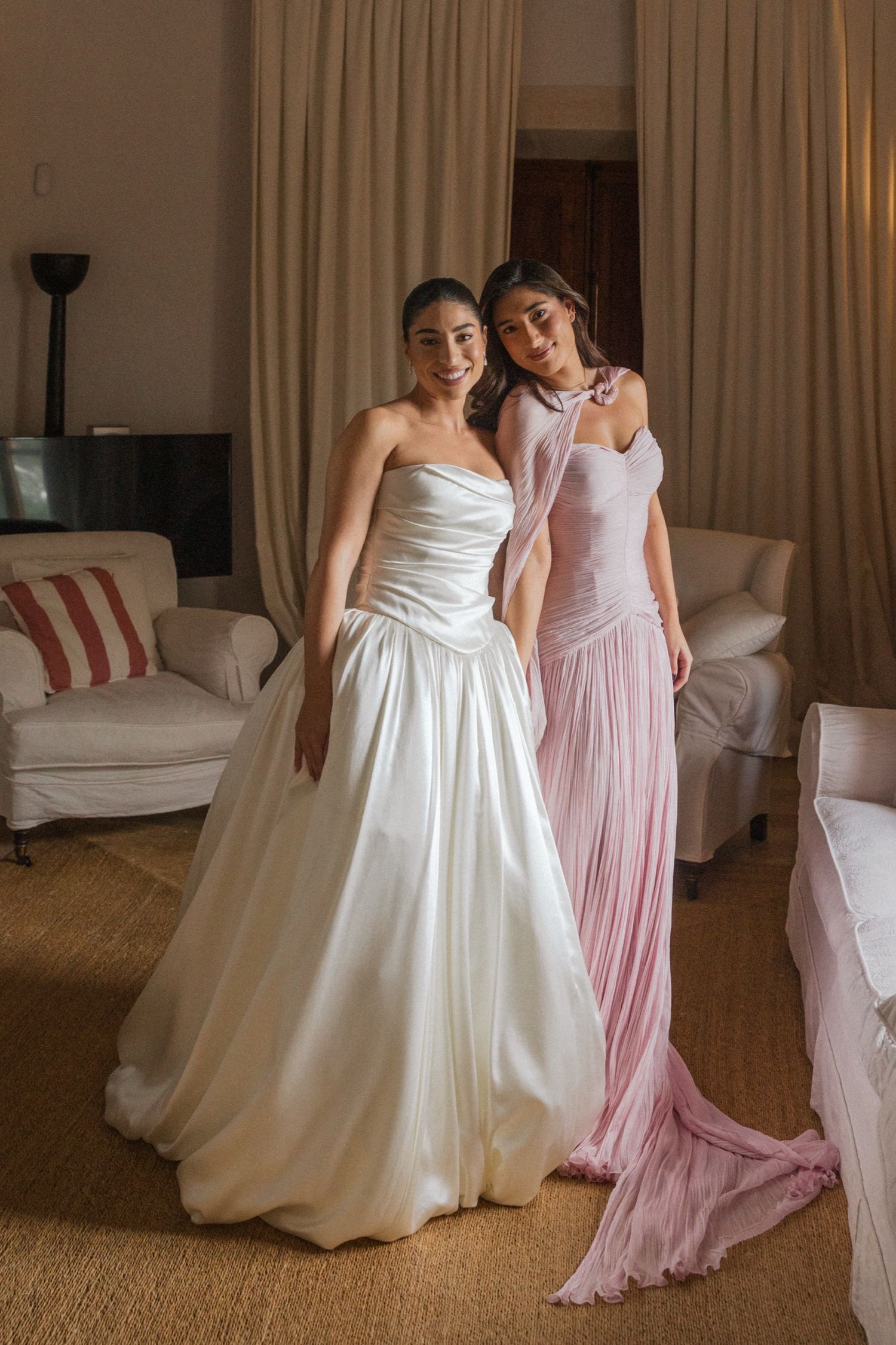 Two people in formal gowns, one in white and one in pink, pose together in an elegant room with neutral decor.
