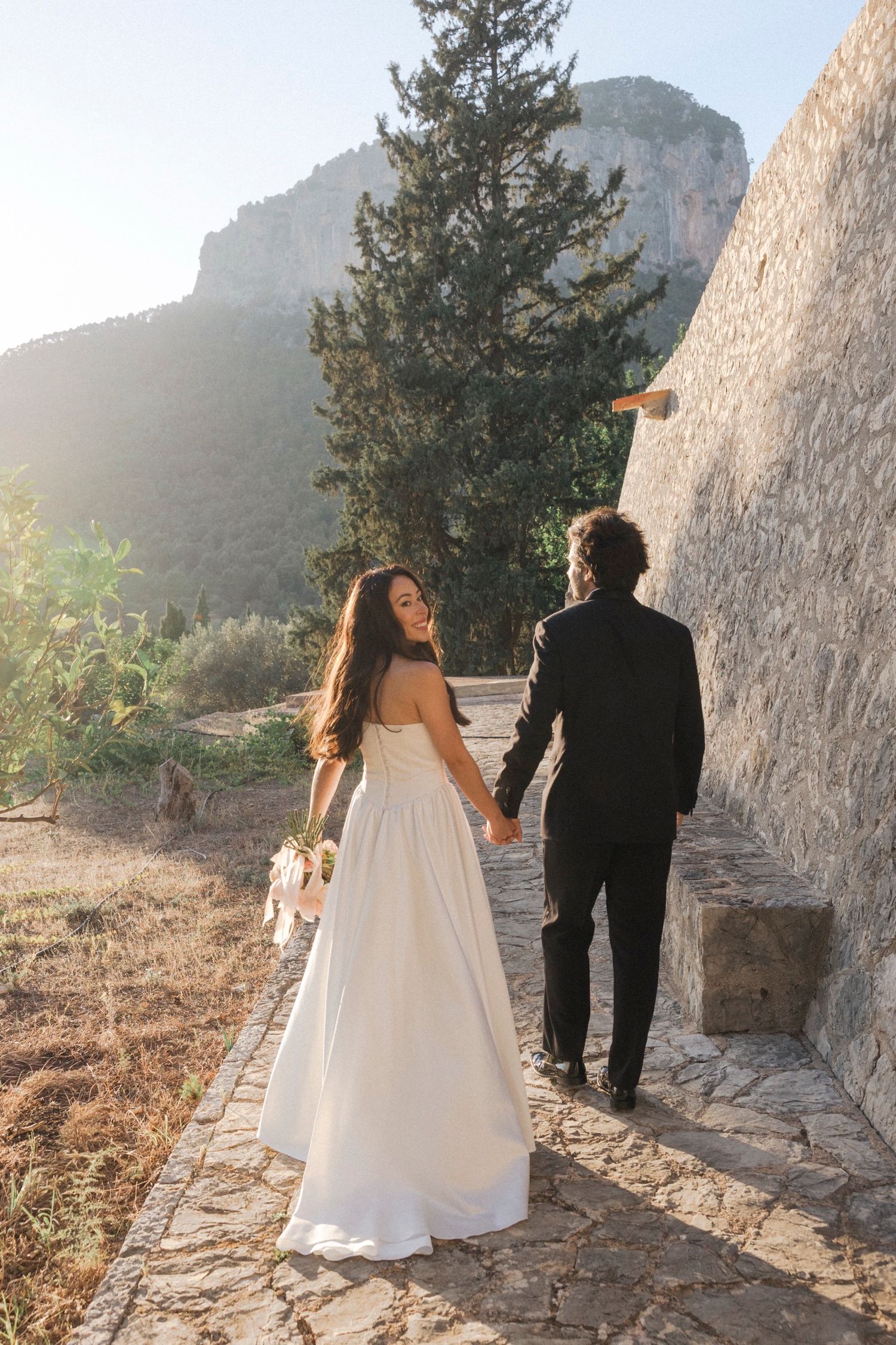 A couple in wedding attire walks along a stone path with mountains and cliffs in the scenic backdrop.