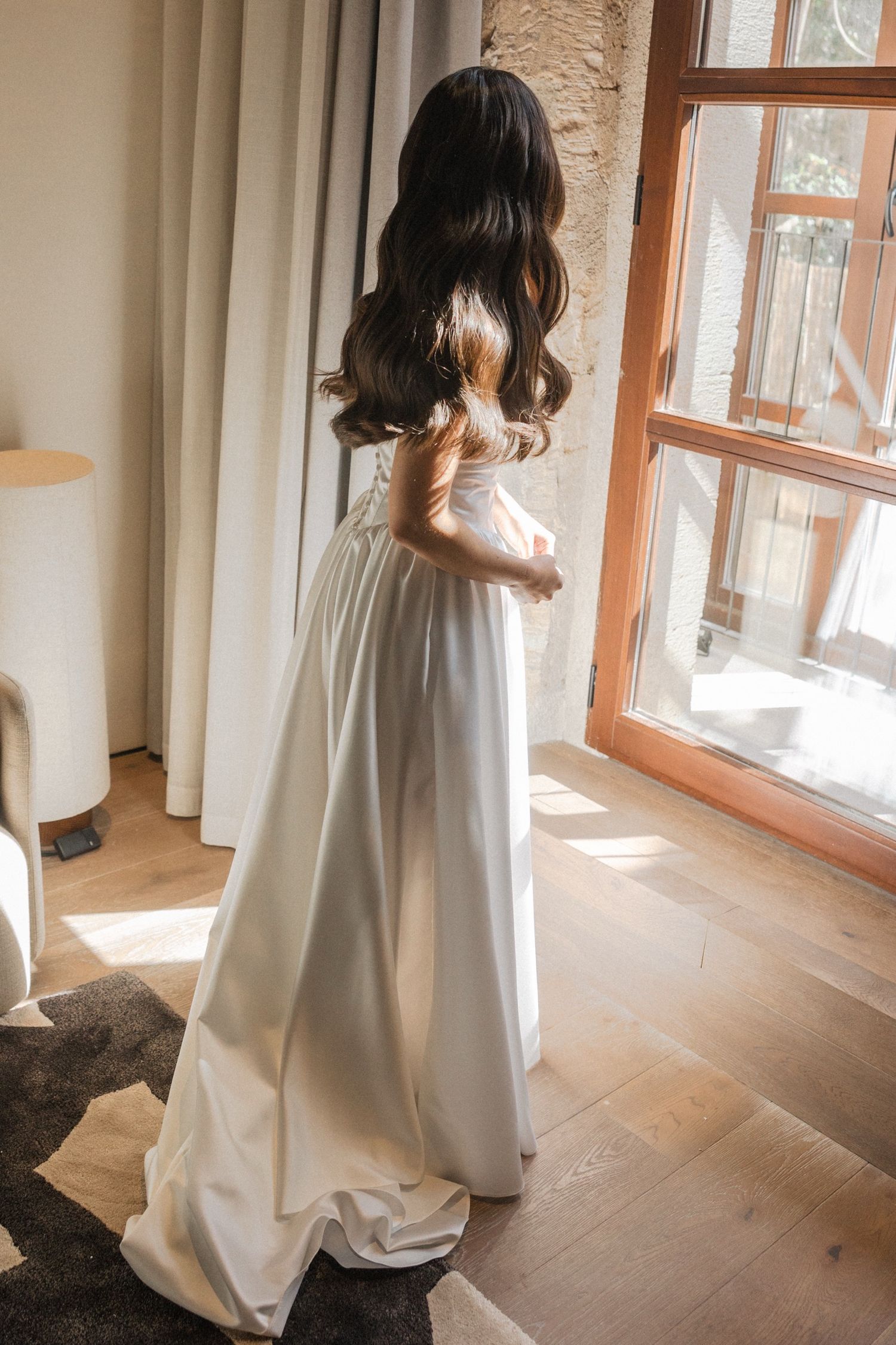 A person in an elegant white flowing dress stands near a sunny window in a minimalist room with wooden floors.