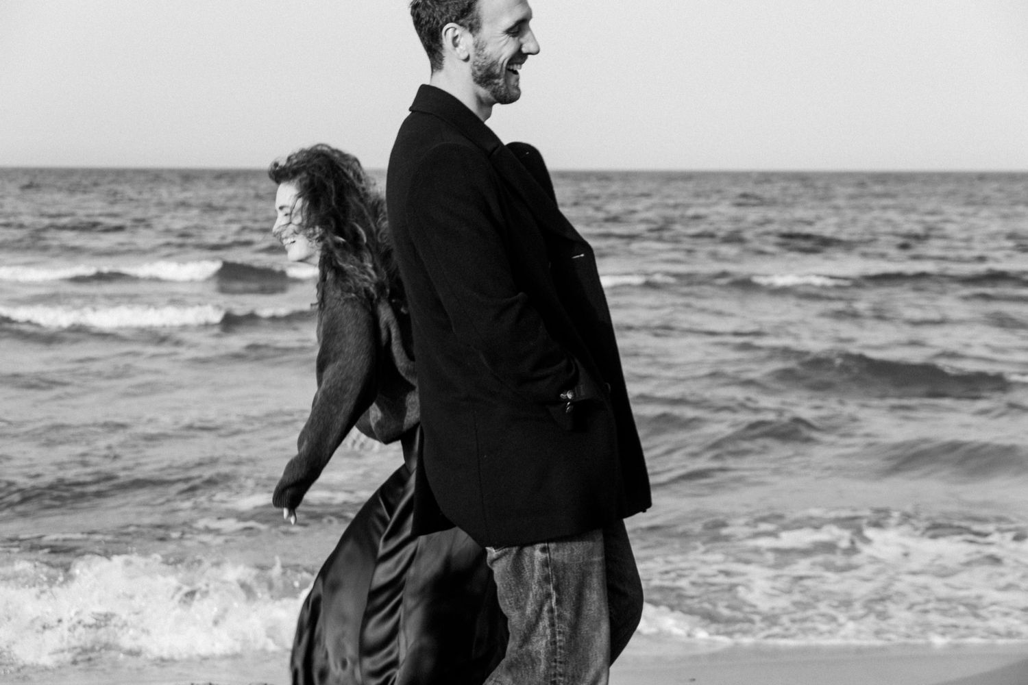 A couple in dark clothing walks along a scenic beach shoreline as waves crash behind them.