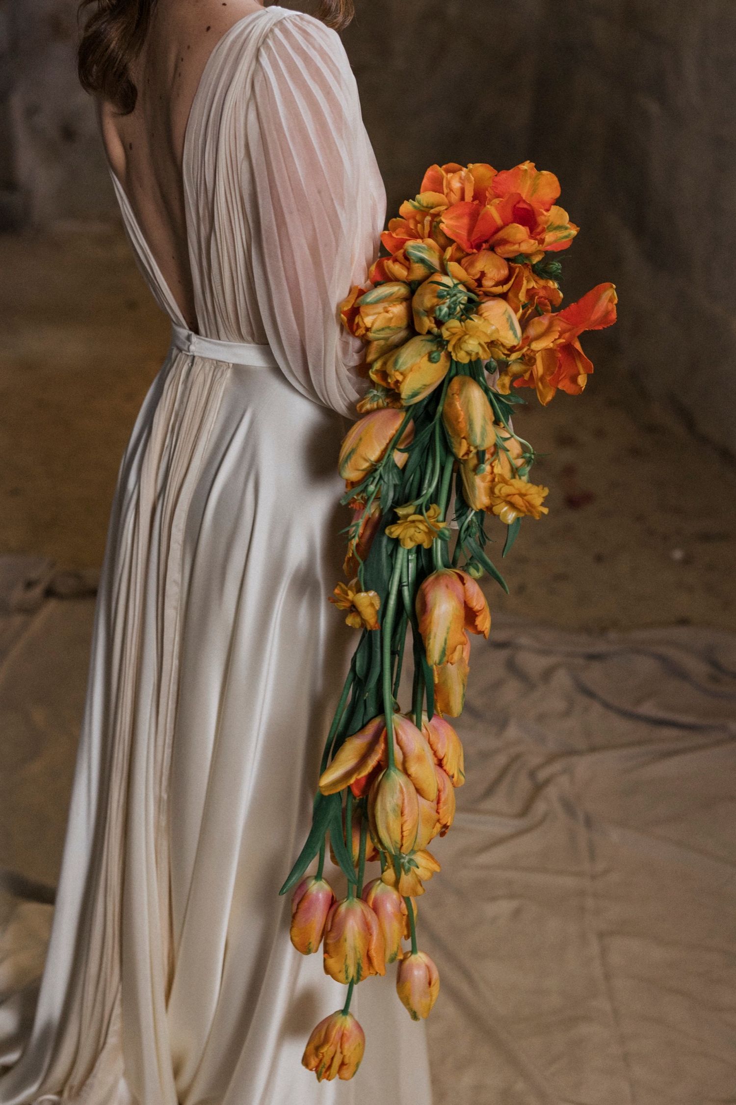 A cascading orange and yellow tulip bouquet adorns the side of a flowing white dress in an artistic fashion shoot.
