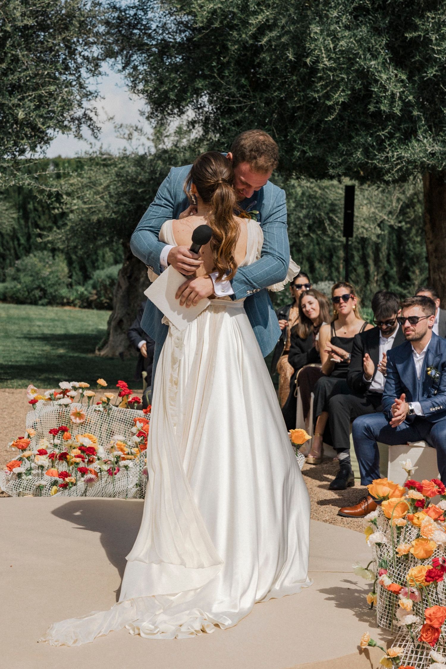 Outdoor wedding ceremony with bright orange flowers decorating the aisle and guests seated under trees.