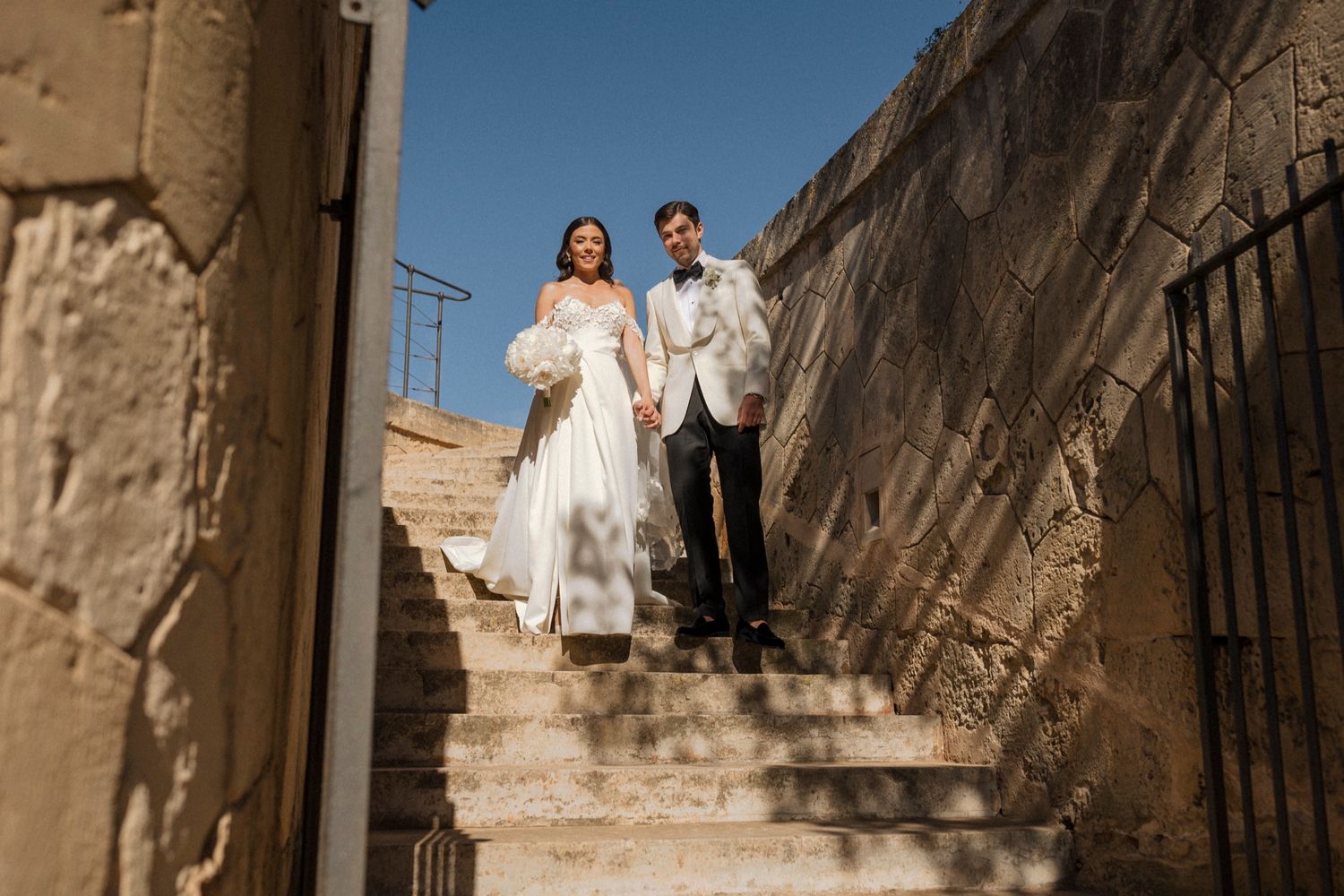 Bride and groom walk down ancient stone steps holding hands on their wedding day under a clear blue sky.