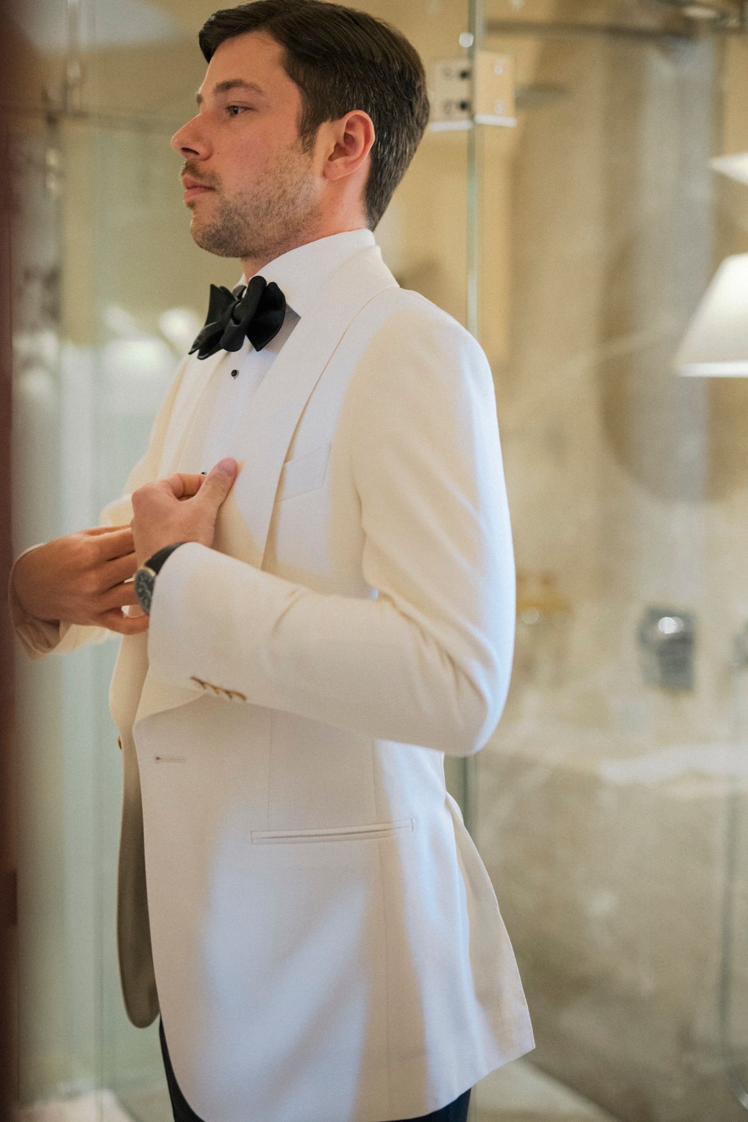Elegant man in white tuxedo jacket and black bow tie adjusting his suit in a mirror.