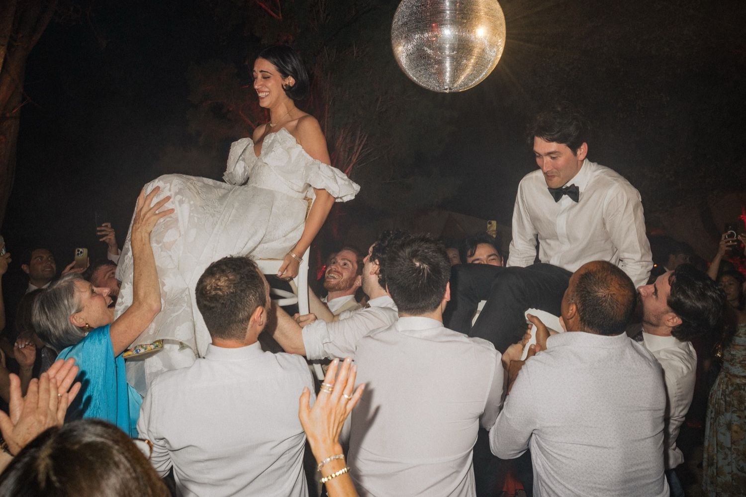 Wedding party dances and celebrates on a crowded dance floor under disco ball lighting at a reception.