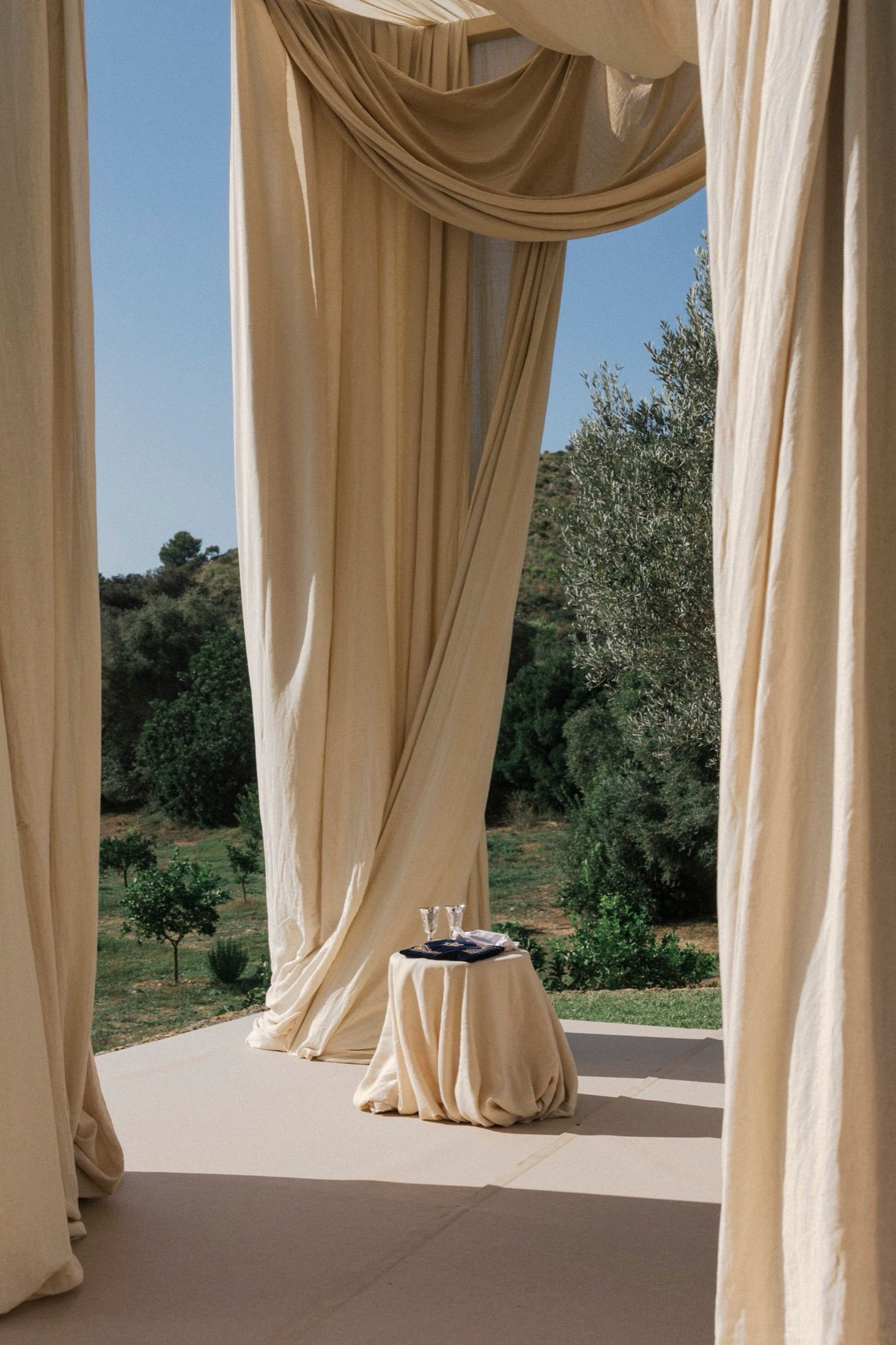 Elegant cream-colored drapes frame an outdoor terrace with a small round table against a scenic natural landscape.
