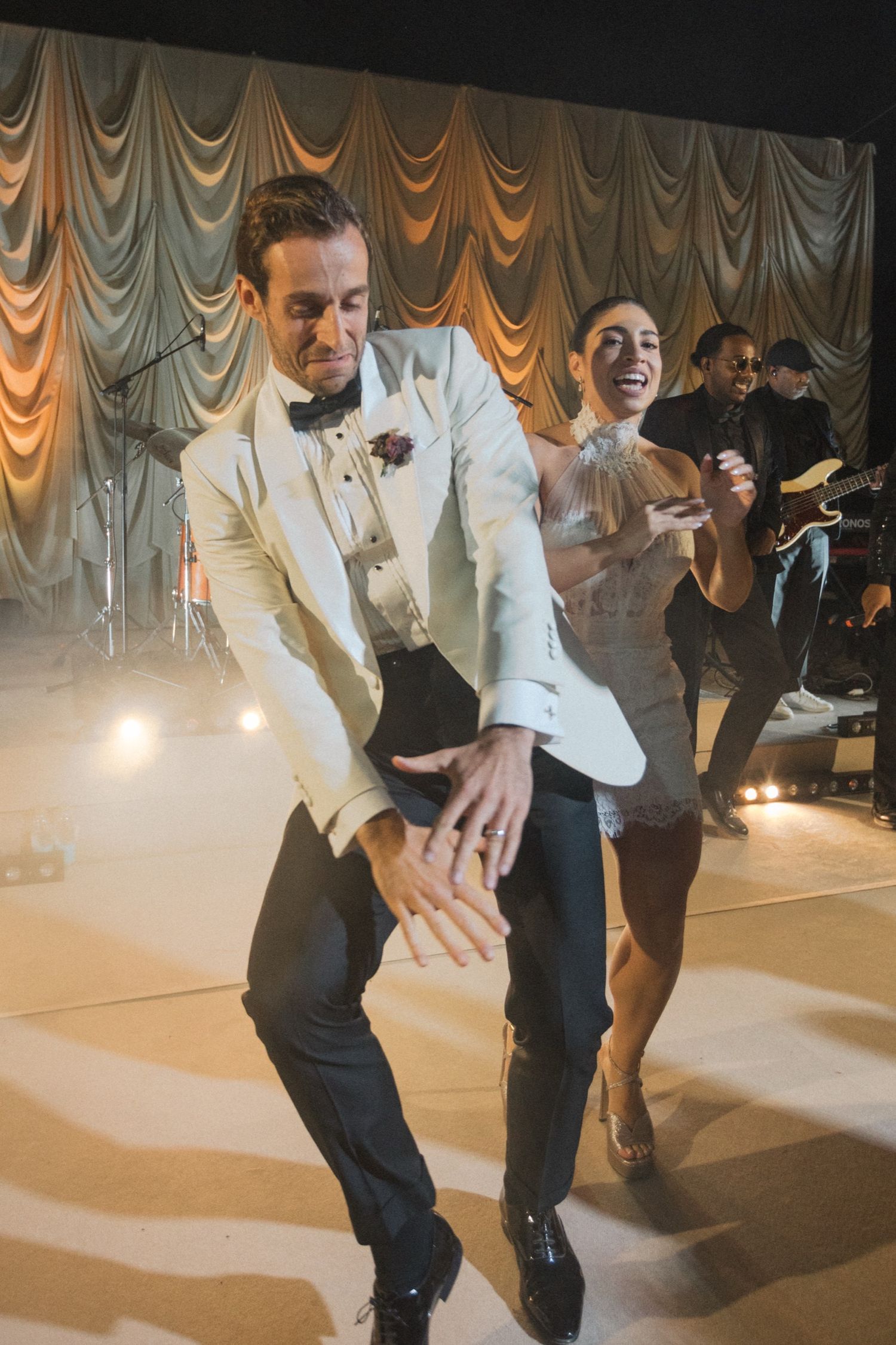 Guests dancing energetically at a wedding reception with elegant draped curtains and stage lighting in the background.