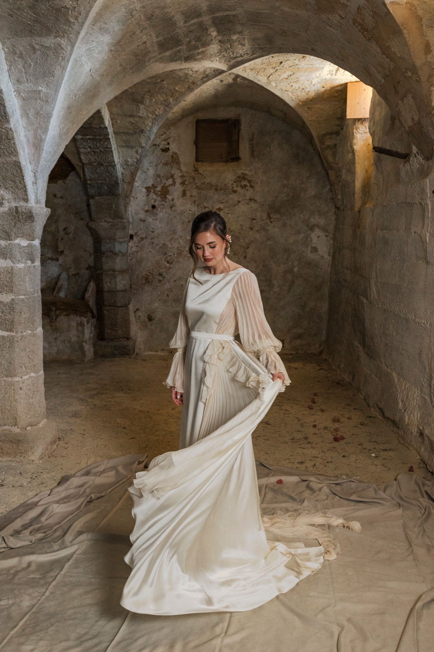 A figure in a flowing white vintage dress poses gracefully in a dimly lit stone cellar with arched vaults and columns.