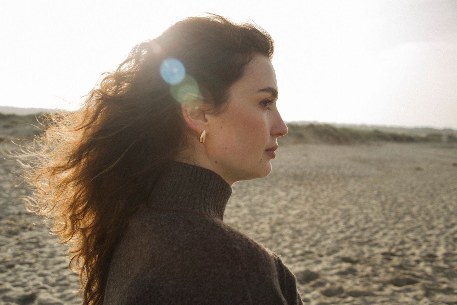 A woman with windswept hair gazes into the distance at sunset on a rocky beach.