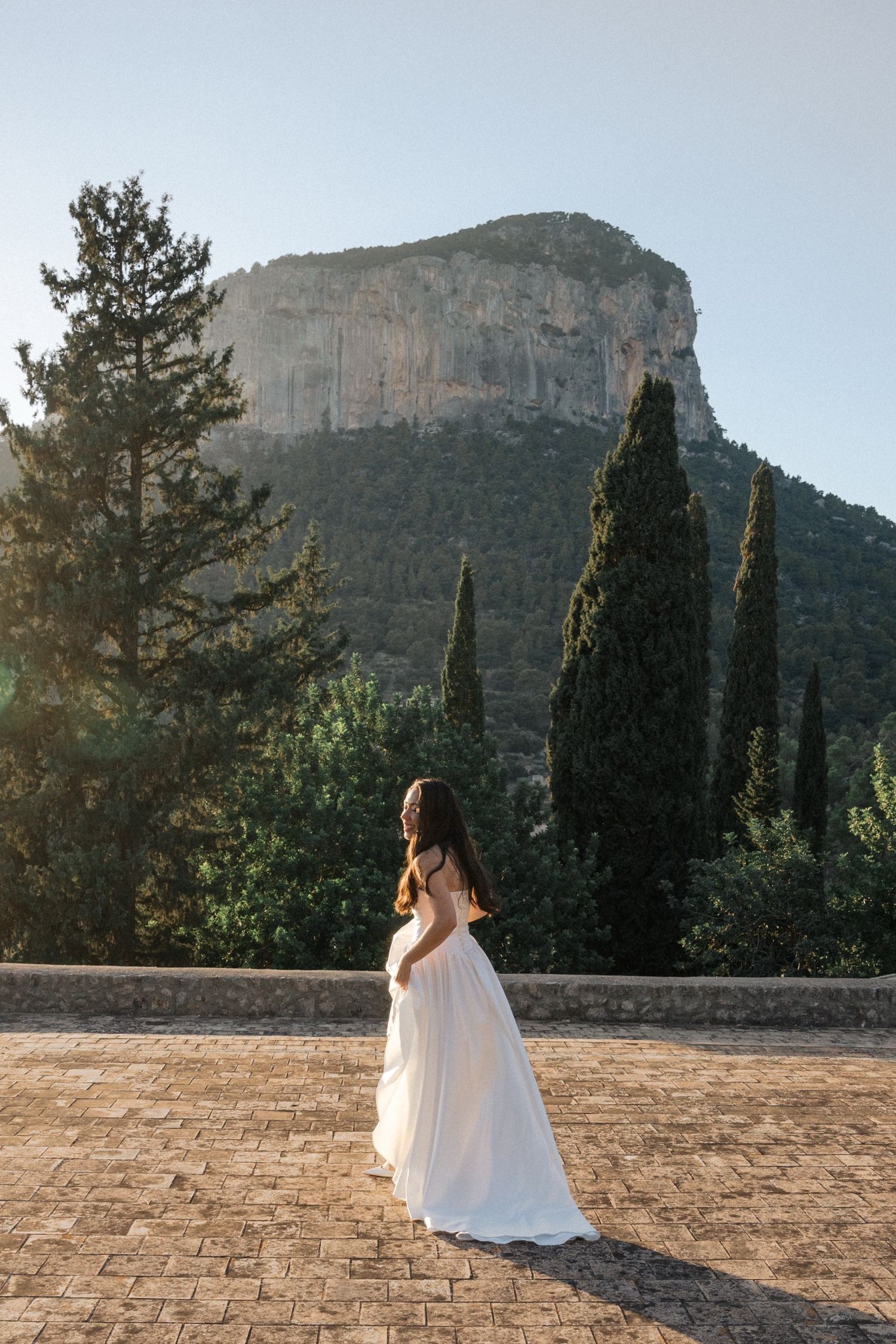 A person in a white dress stands against a backdrop of majestic mountains and cypress trees in a Mediterranean landscape.