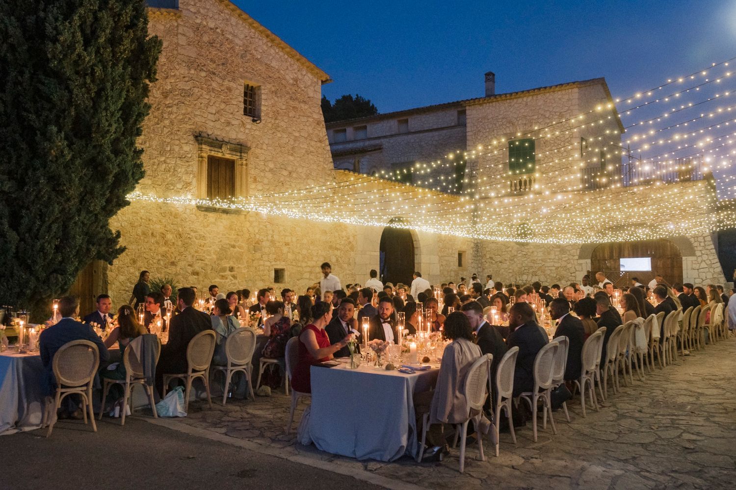 Long rows of tables and chairs set up for an outdoor evening event under string lights in a rustic stone courtyard.