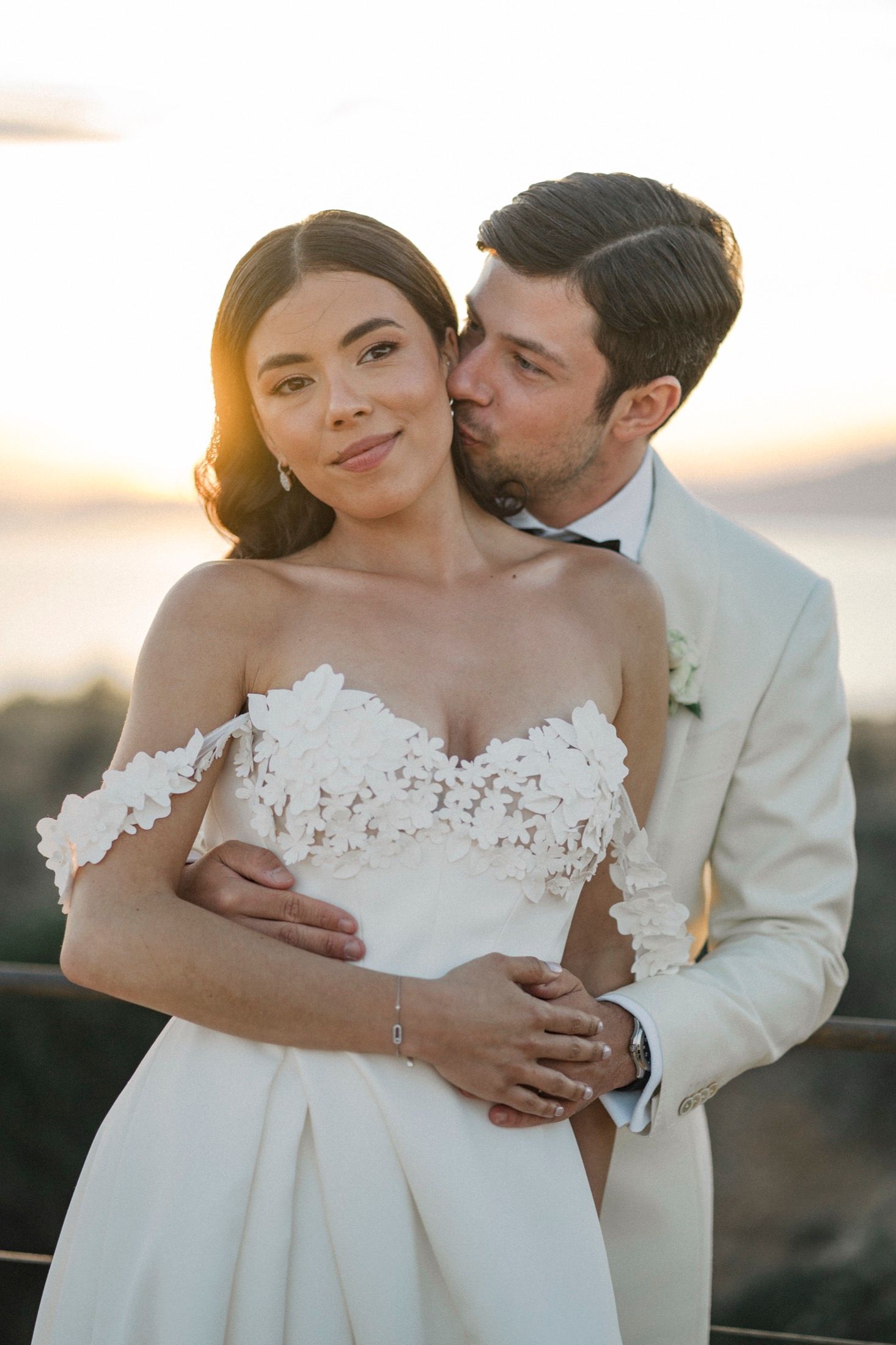 Bride in floral off-shoulder gown embraced by groom in ivory suit at golden hour sunset wedding.