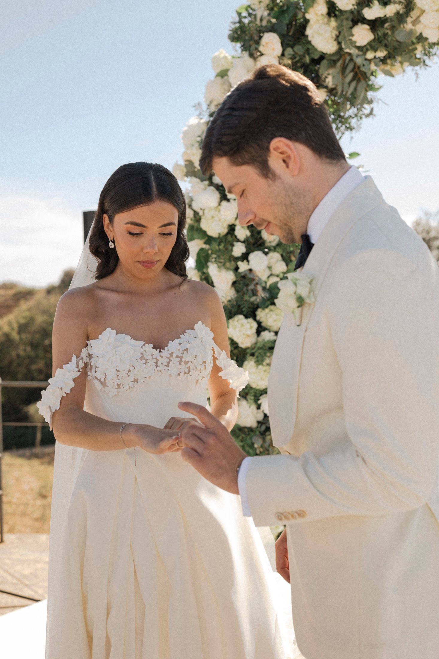 Groom in white suit placing wedding ring on bride's finger at outdoor ceremony with white floral arch.