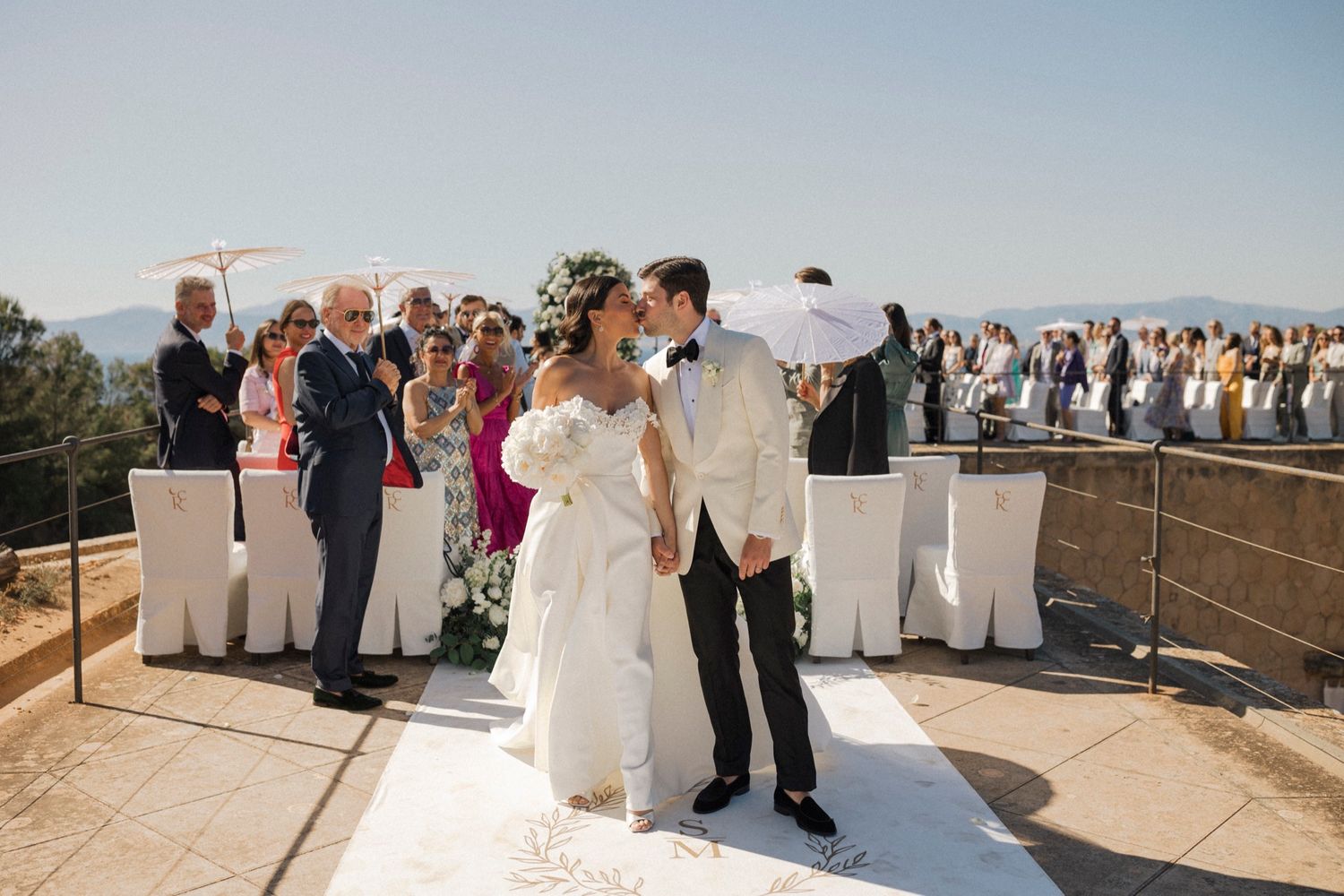 Bride and groom walk down outdoor aisle after wedding ceremony with guests and scenic views behind them.