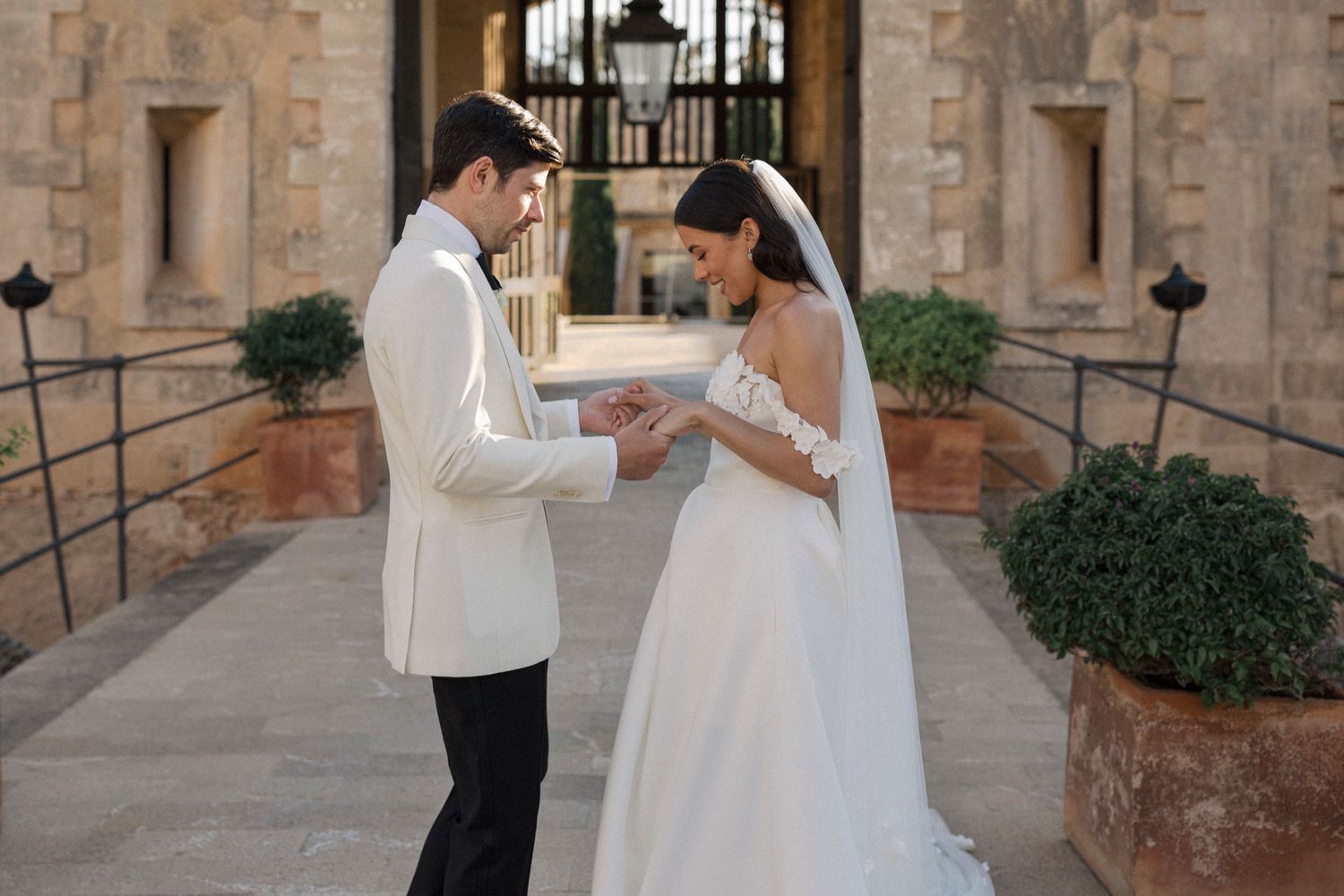 Bride and groom exchanging rings outdoors at a stone venue, groom in white tuxedo jacket, bride in elegant white gown.