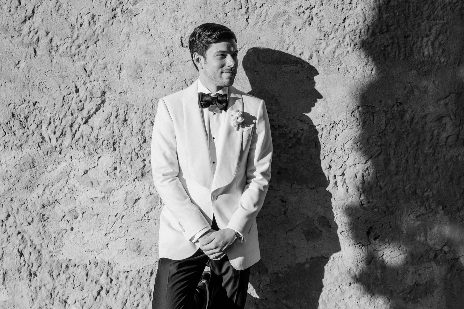 Groom in white tuxedo jacket and bow tie poses against textured stone wall in elegant black and white wedding portrait.