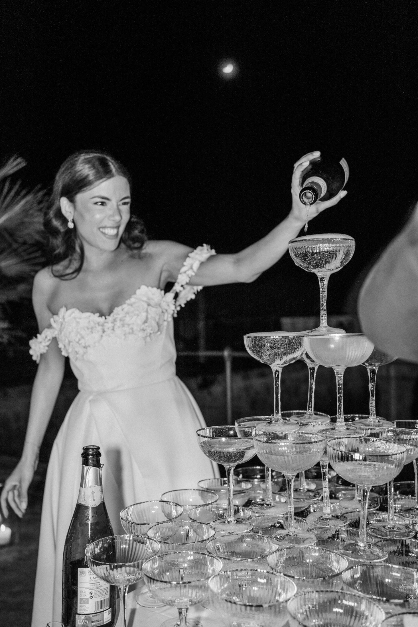 Bride in white gown pouring champagne into a towering pyramid of coupe glasses at a night wedding reception.