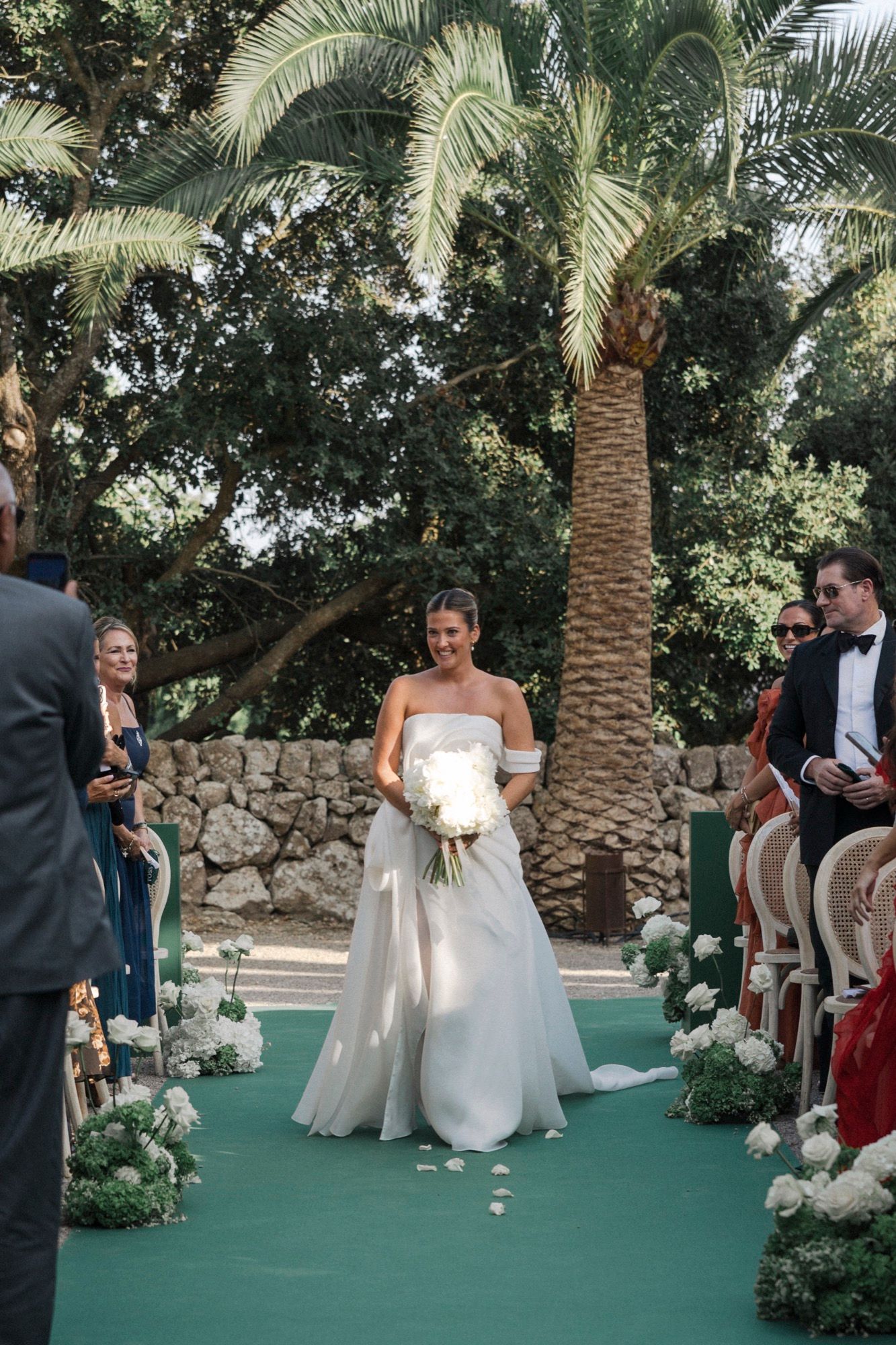Outdoor wedding ceremony with green carpet aisle and palm trees in a Mediterranean setting.
