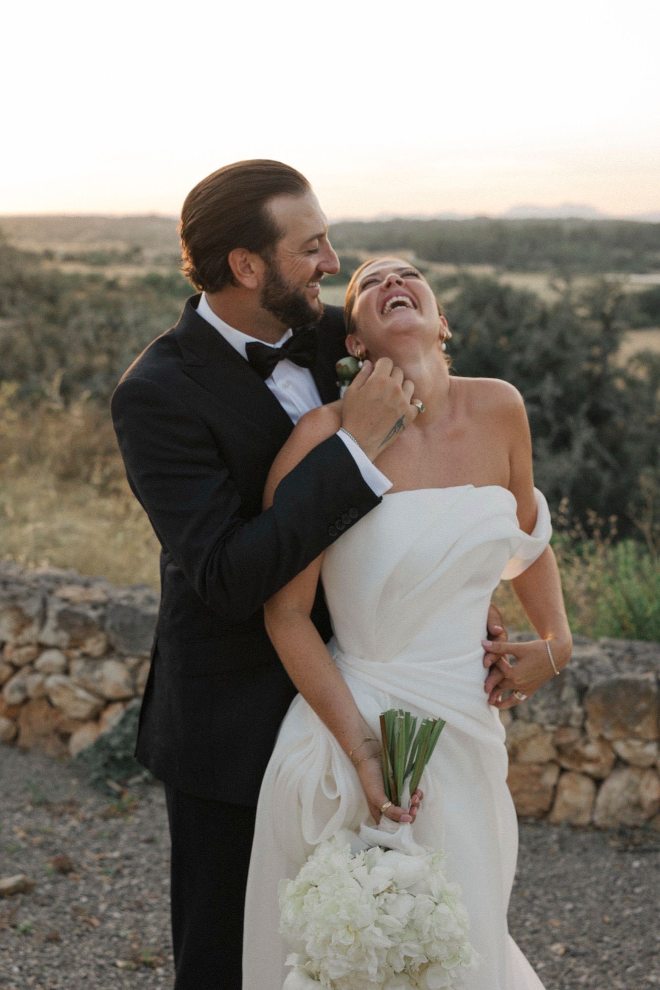 Joyful wedding couple shares romantic moment while holding white bouquet against scenic backdrop.