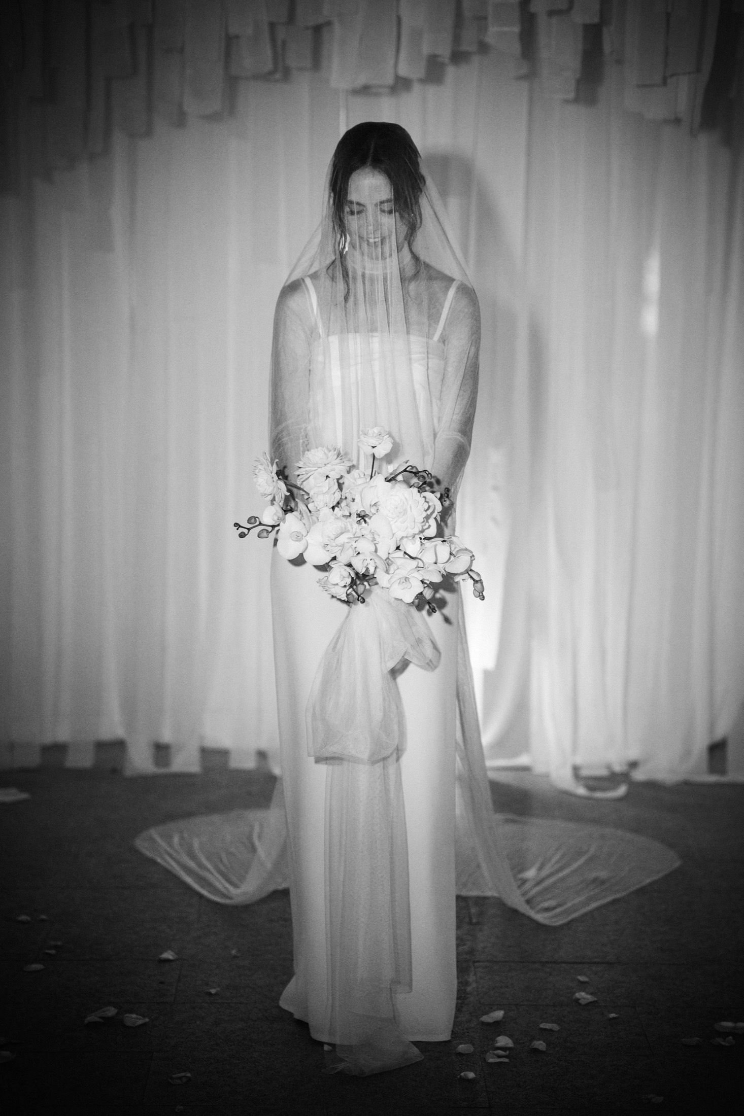 A black and white artistic bridal portrait showing a figure in a flowing wedding gown holding a bouquet of flowers.