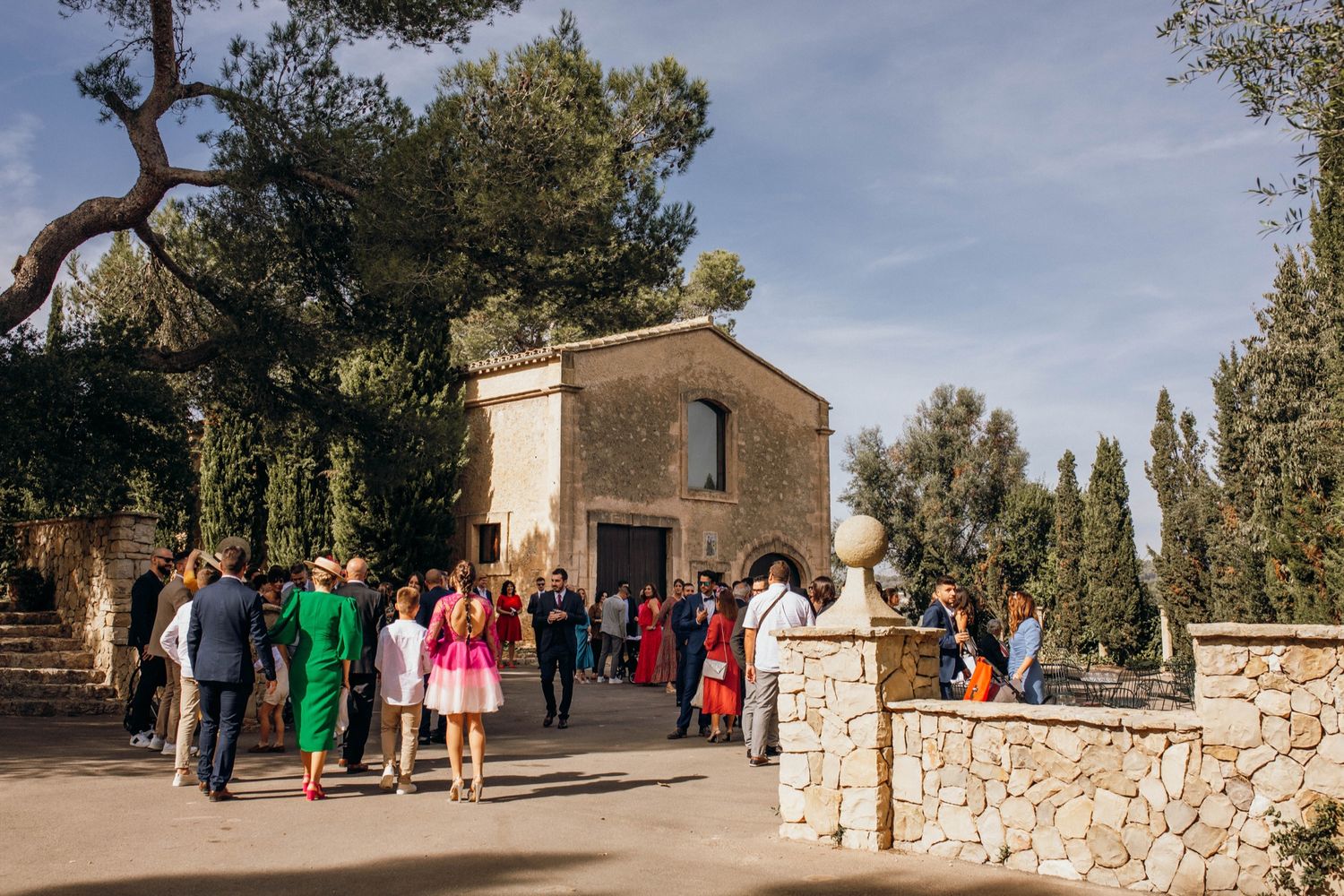 People walking along a stone-walled path near rustic buildings in a Mediterranean countryside setting on a sunny day.