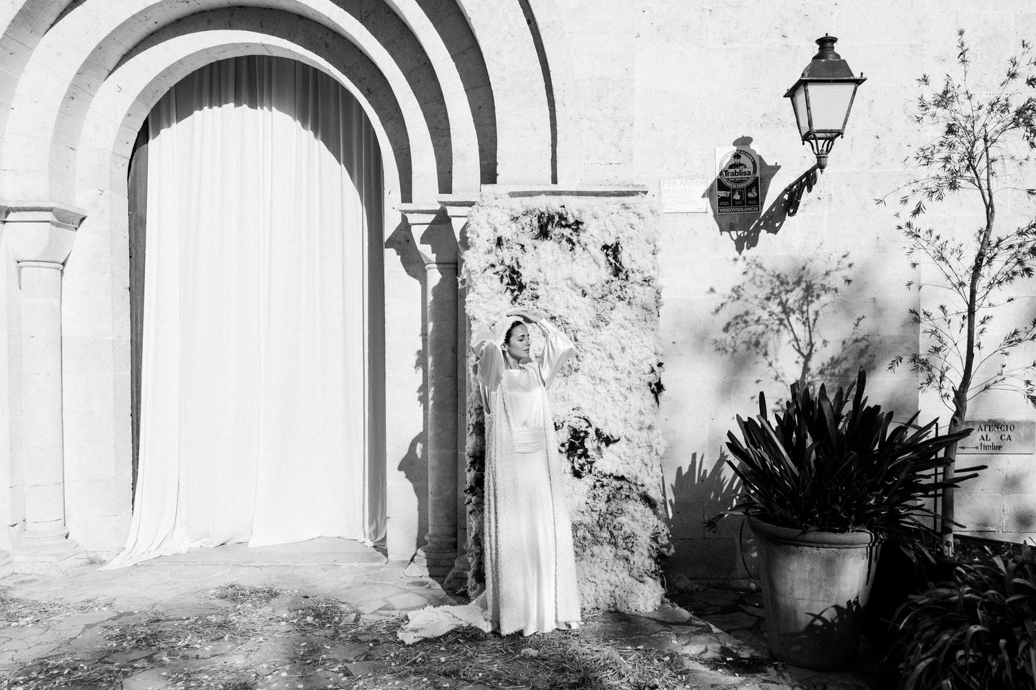 Artistic black and white photo of a figure in flowing white dress against stone archways and garden foliage.