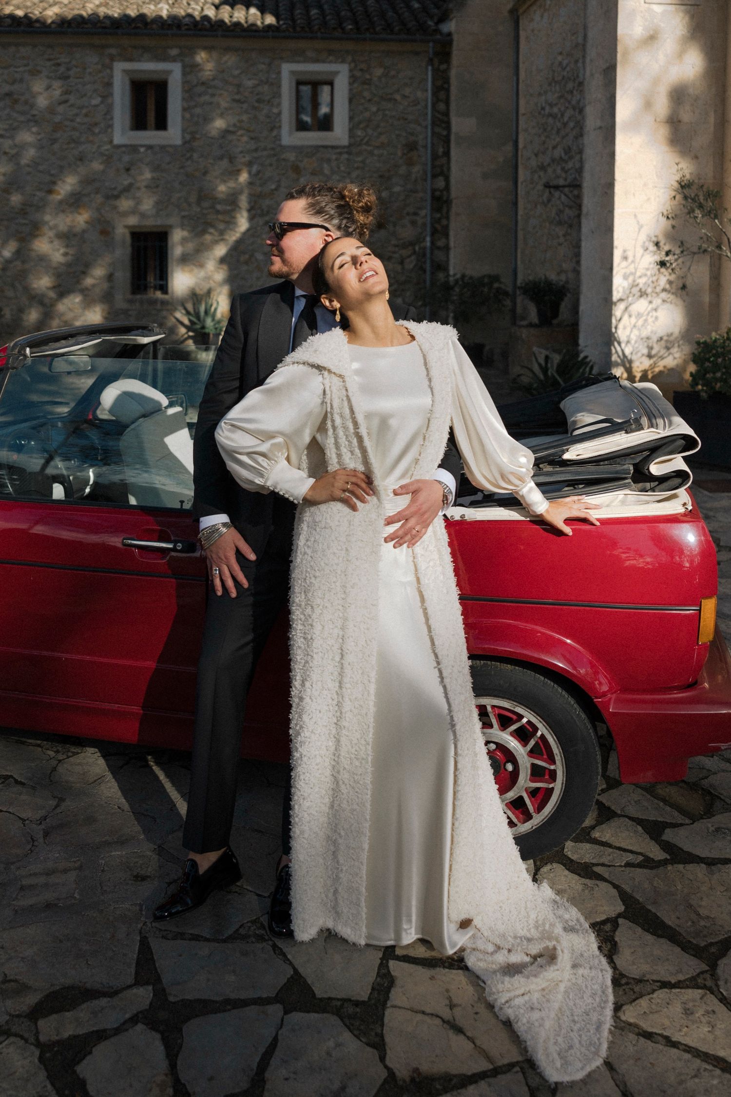 Bride in vintage-style dress posing with classic red sports car against stone building backdrop.