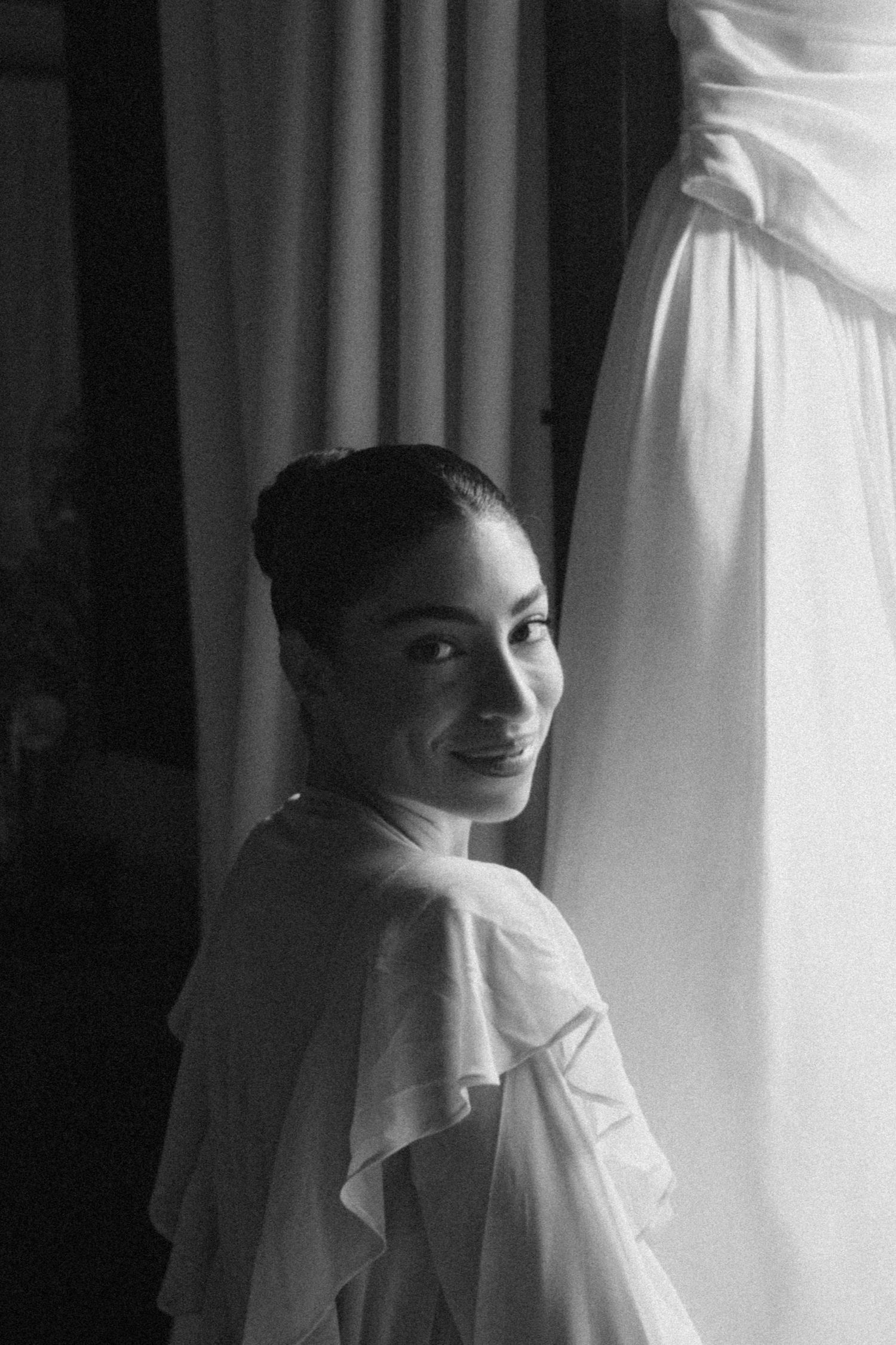 Black and white portrait of a person in elegant white attire looking over their shoulder near a window with soft lighting.