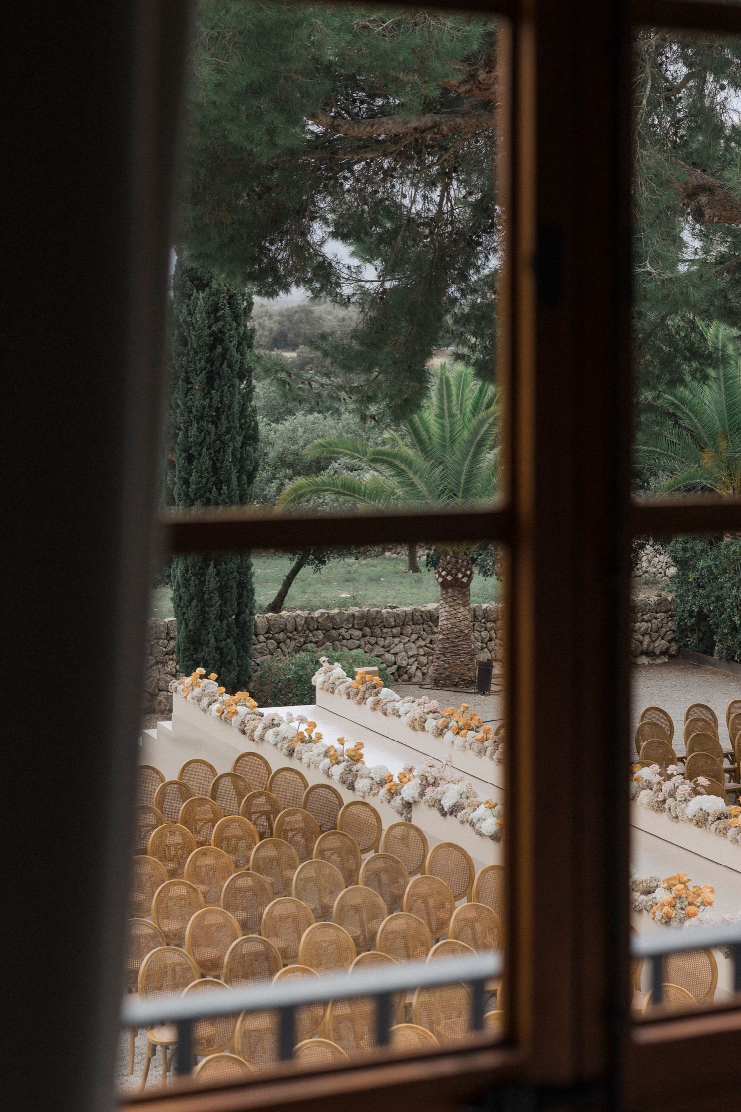 View through a window showing rows of wooden chairs set up for an outdoor wedding ceremony.