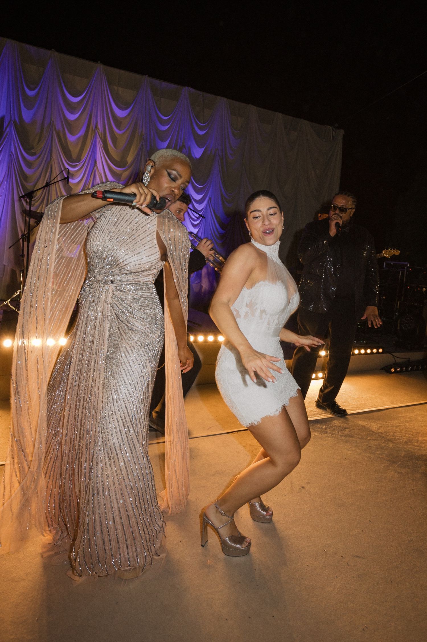 Two people dancing on stage in glamorous evening wear with purple curtains and stage lighting in the background.