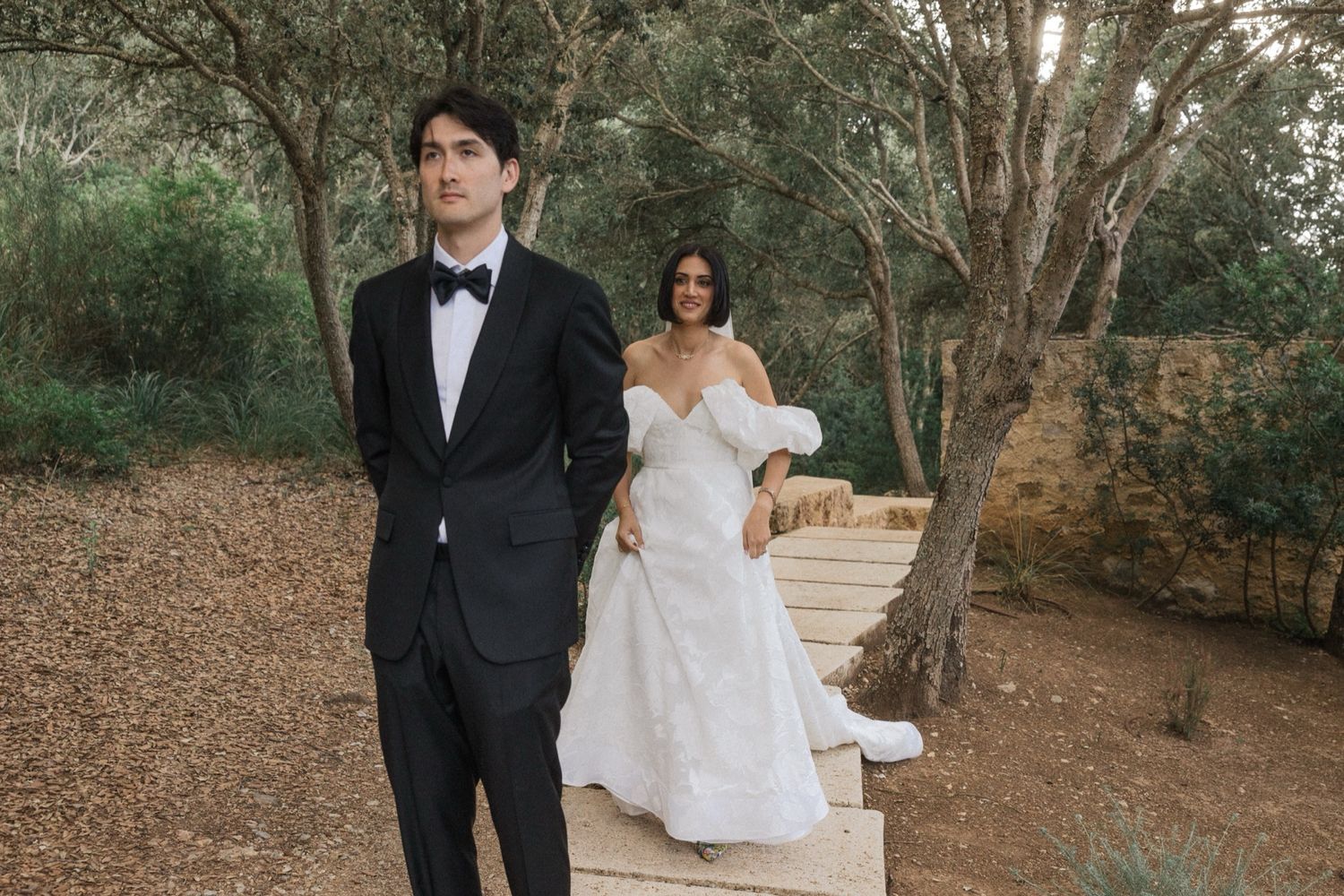 A couple in formal wedding attire stands on a path surrounded by trees during their outdoor ceremony.