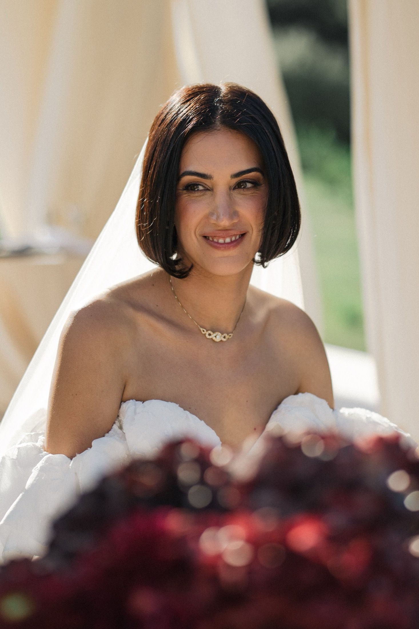 A bride in a white wedding dress smiles while standing behind dark red flowers during an outdoor ceremony.