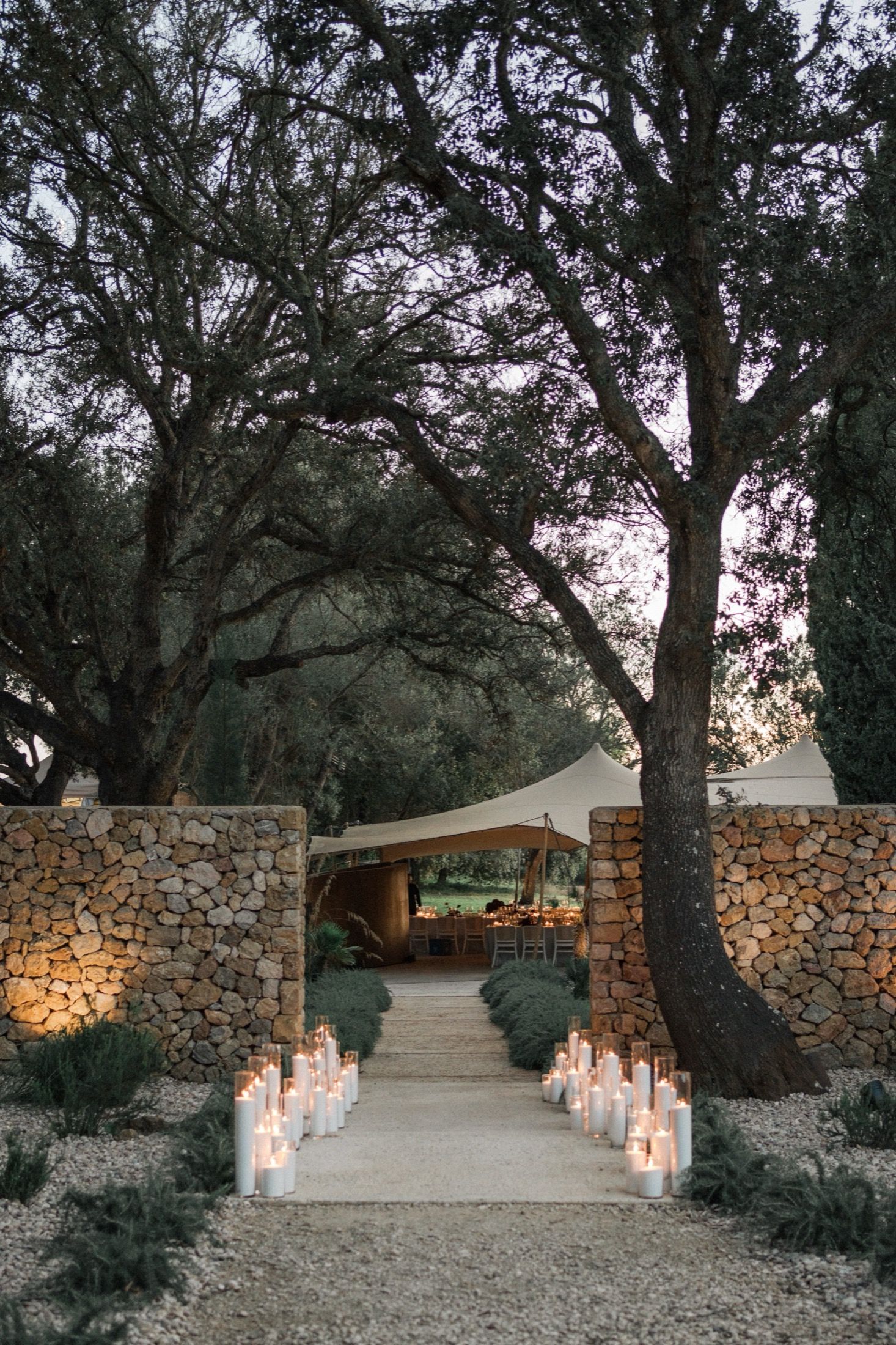 A romantic pathway lined with glowing candles leads through stone walls to an illuminated venue under tall trees at dusk.