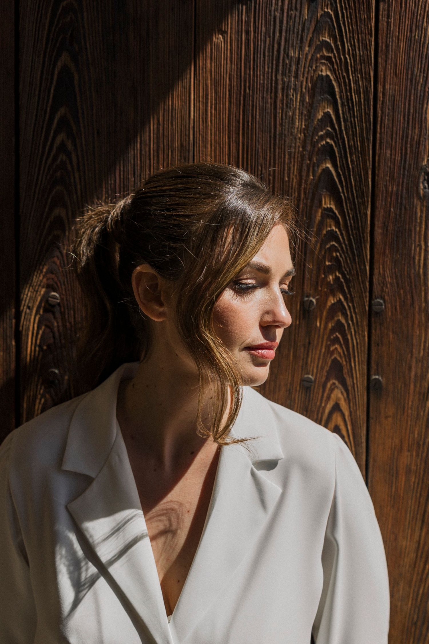 Fashion model in white blazer poses against dark wooden wall with dramatic lighting and shadows.